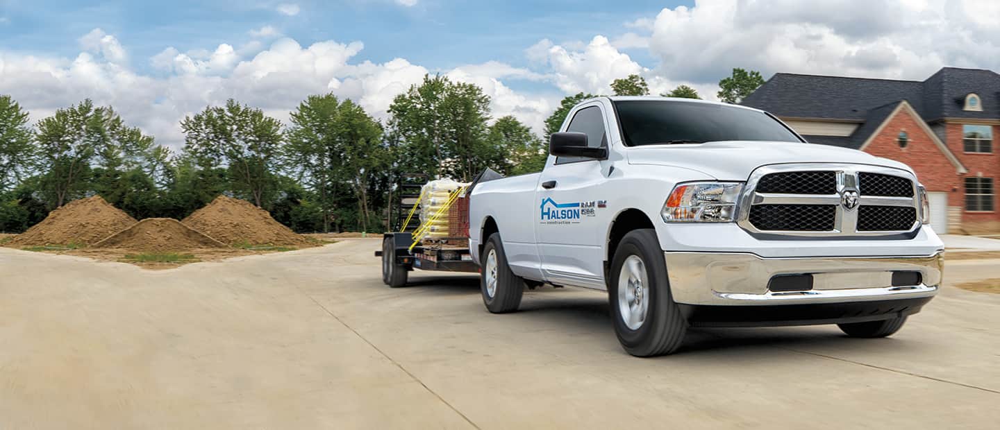 A white 2023 Ram 1500 Classic Tradesman Regular Cab with construction company signage on its passenger-side door, towing building materials on a flatbed trailer.