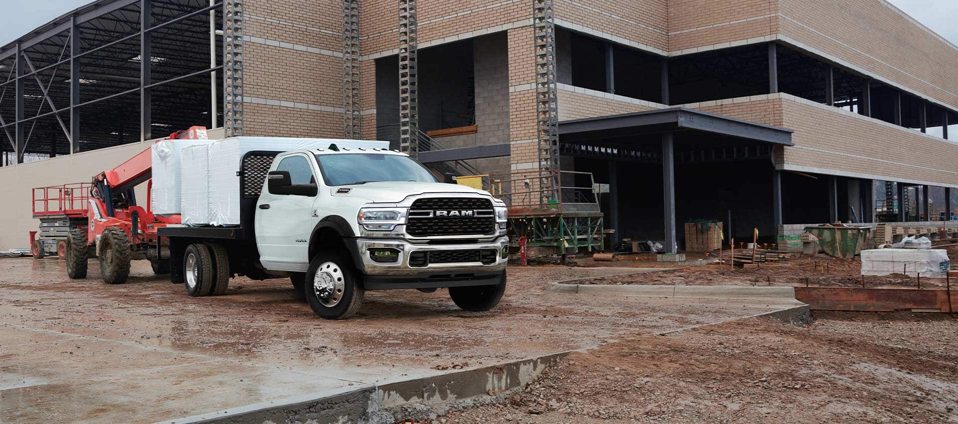 A white 2024 Ram Chassis Cab with a platform upfit loaded with building materials, parked at a commercial construction site.