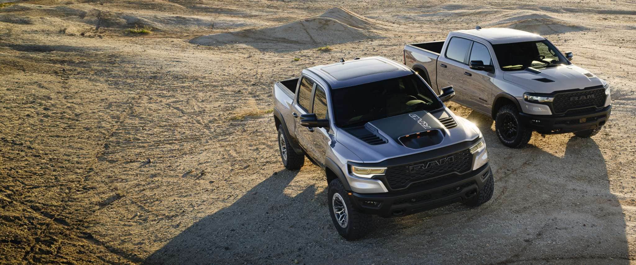 Two Ram Brand Off-Road Trucks parked off-road in the desert: on the left, a silver 2025 Ram 1500 RHO Crew Cab and on the right a silver 2025 Ram 1500 Rebel X Crew Cab.