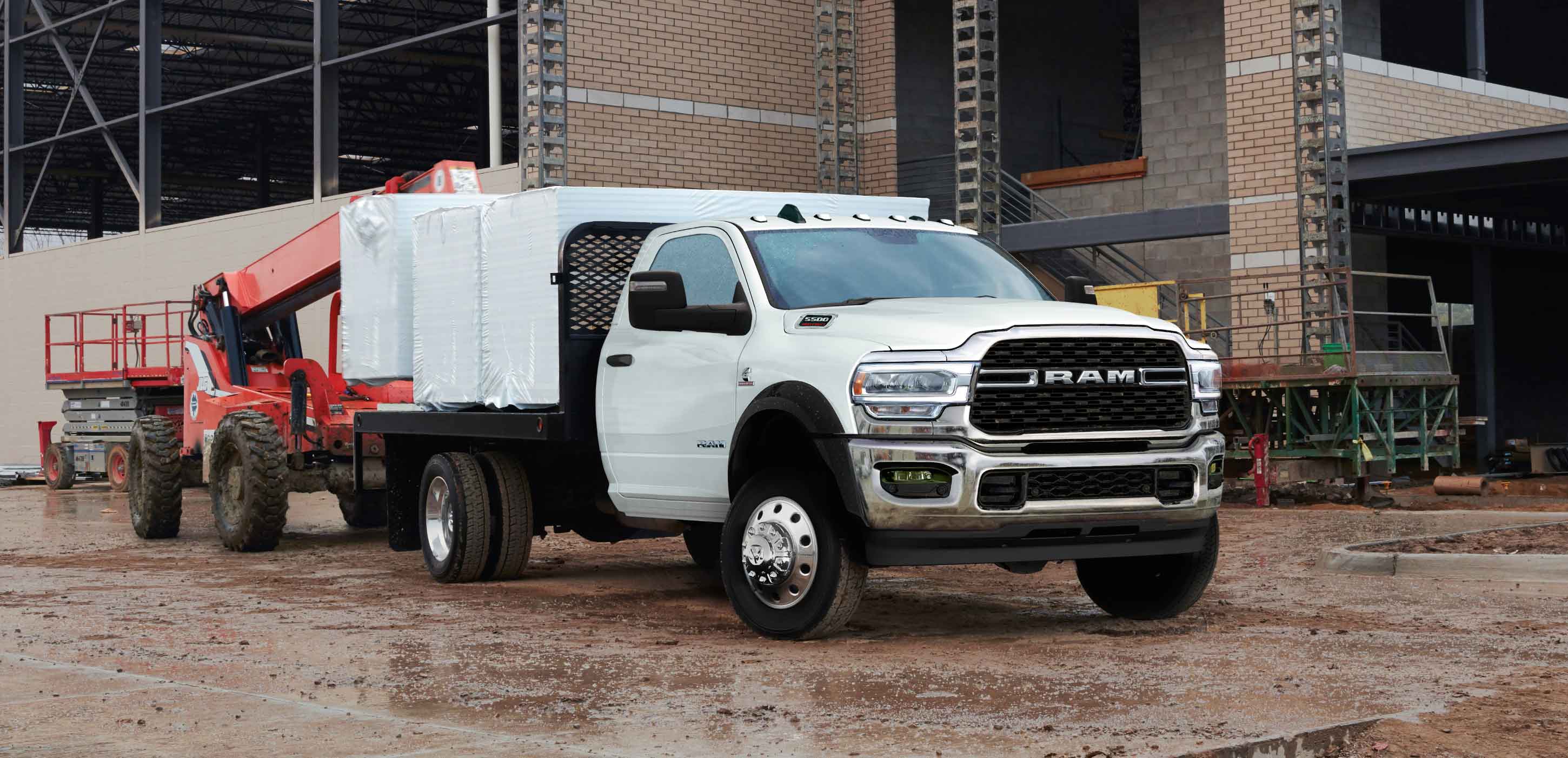Display A white 2024 Ram 5500 SLT Chassis Cab Regular Cab with a platform upfit loaded with building materials, parked at a commercial construction site.