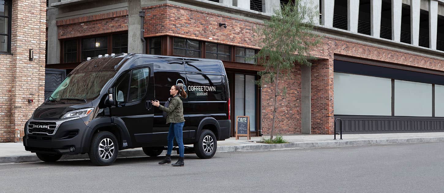 A black 2024 Ram ProMaster 2500 SLT Cargo Van High Roof with a coffee company's signage on its driver-side rear panel, parked on a city street beside a cafe.