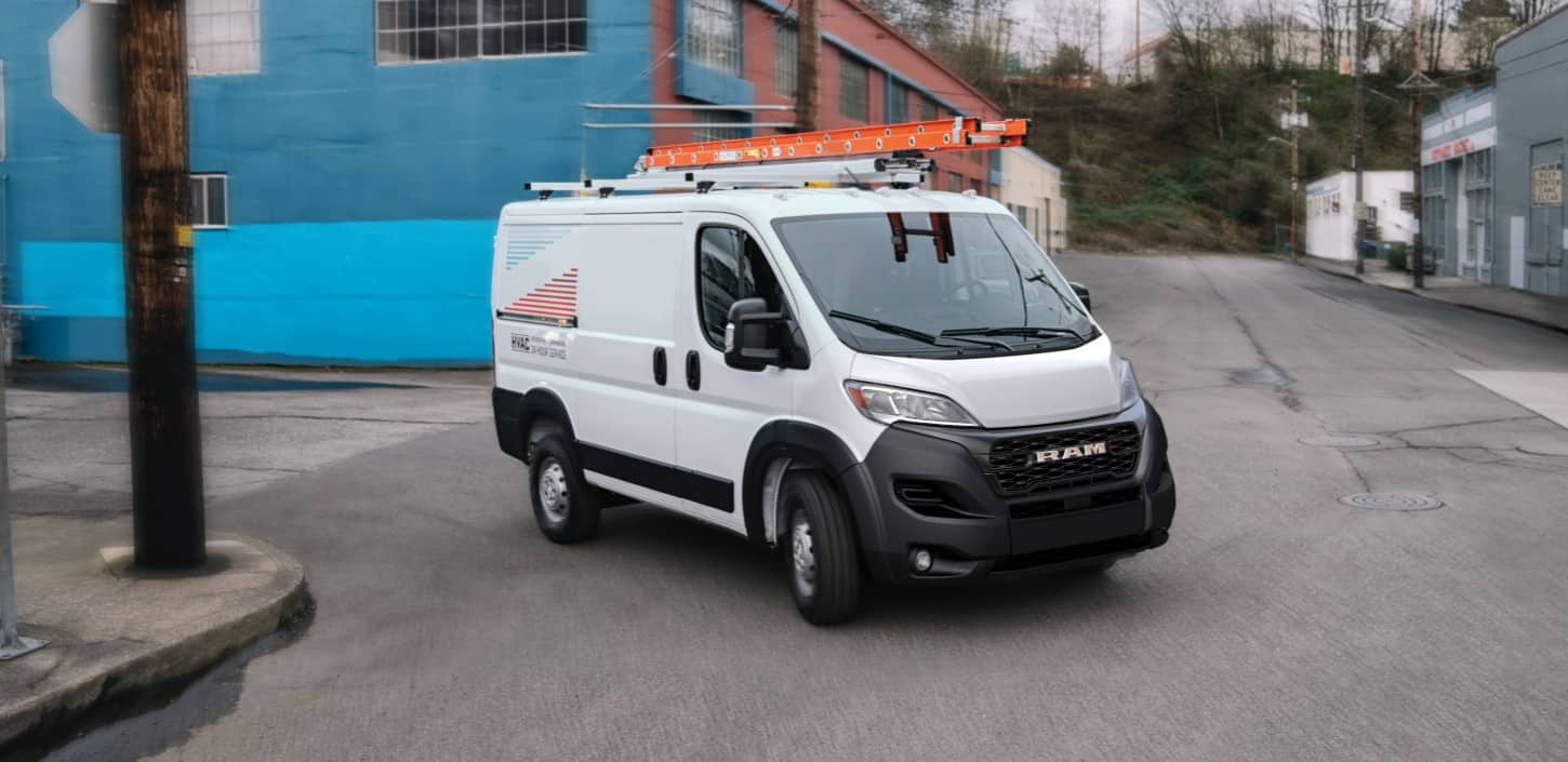 Display A white 2024 Ram ProMaster 1500 Tradesman Cargo Van Standard Roof with the logo for an HVAC company on its side and a ladder on its roof rack making a right turn through an industrial area.