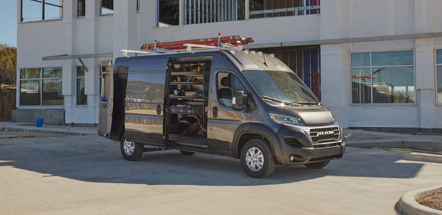 Display A passenger-side profile of a gray 2024 Ram ProMaster SLT Cargo Van High Roof with its sliding side door and rear doors open and a ladder on its roof rack, parked beside a commercial building.