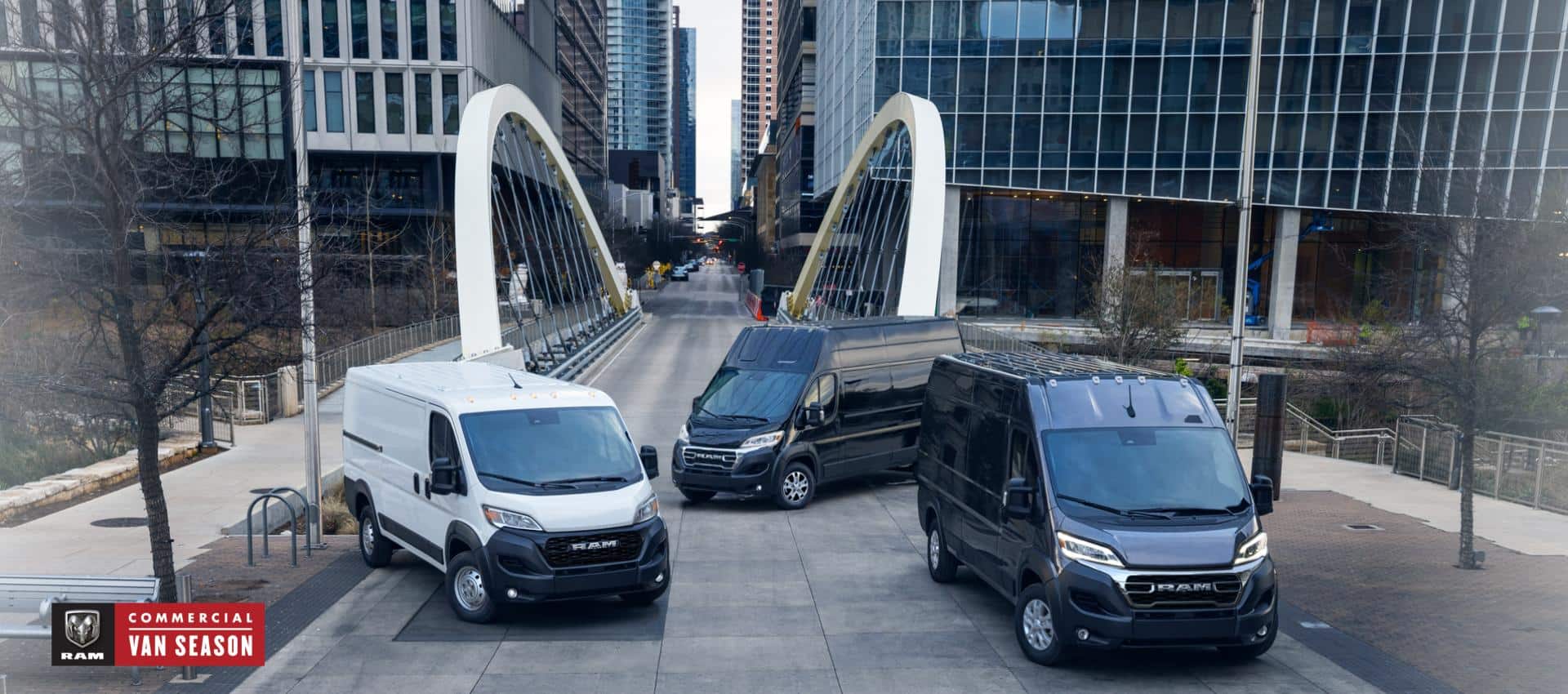 Three 2024 Ram ProMaster models, from left to right: a white Ram ProMaster 1500 Tradesman Standard Roof Cargo Van, a black Ram ProMaster 3500 SLT+ Super High Roof Cargo Van and a black Ram ProMaster 3500 SLT High Roof Cargo Van parked just beyond a bridge in the center of a busy large city. Ram Commercial Van Season.