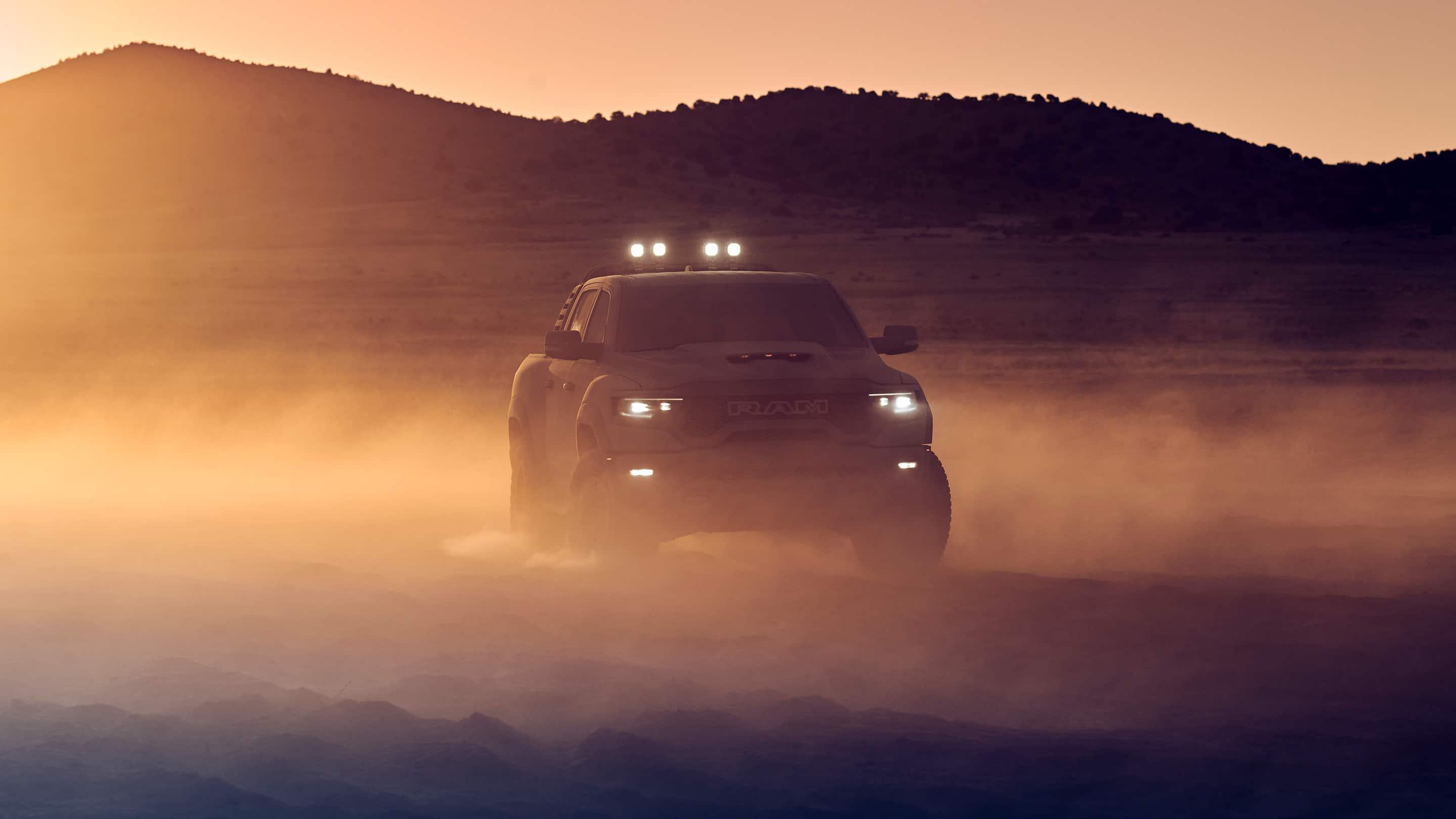 The headlamps of the 2021 Ram 1500 TRX lit as it is driven off-road at sunset.