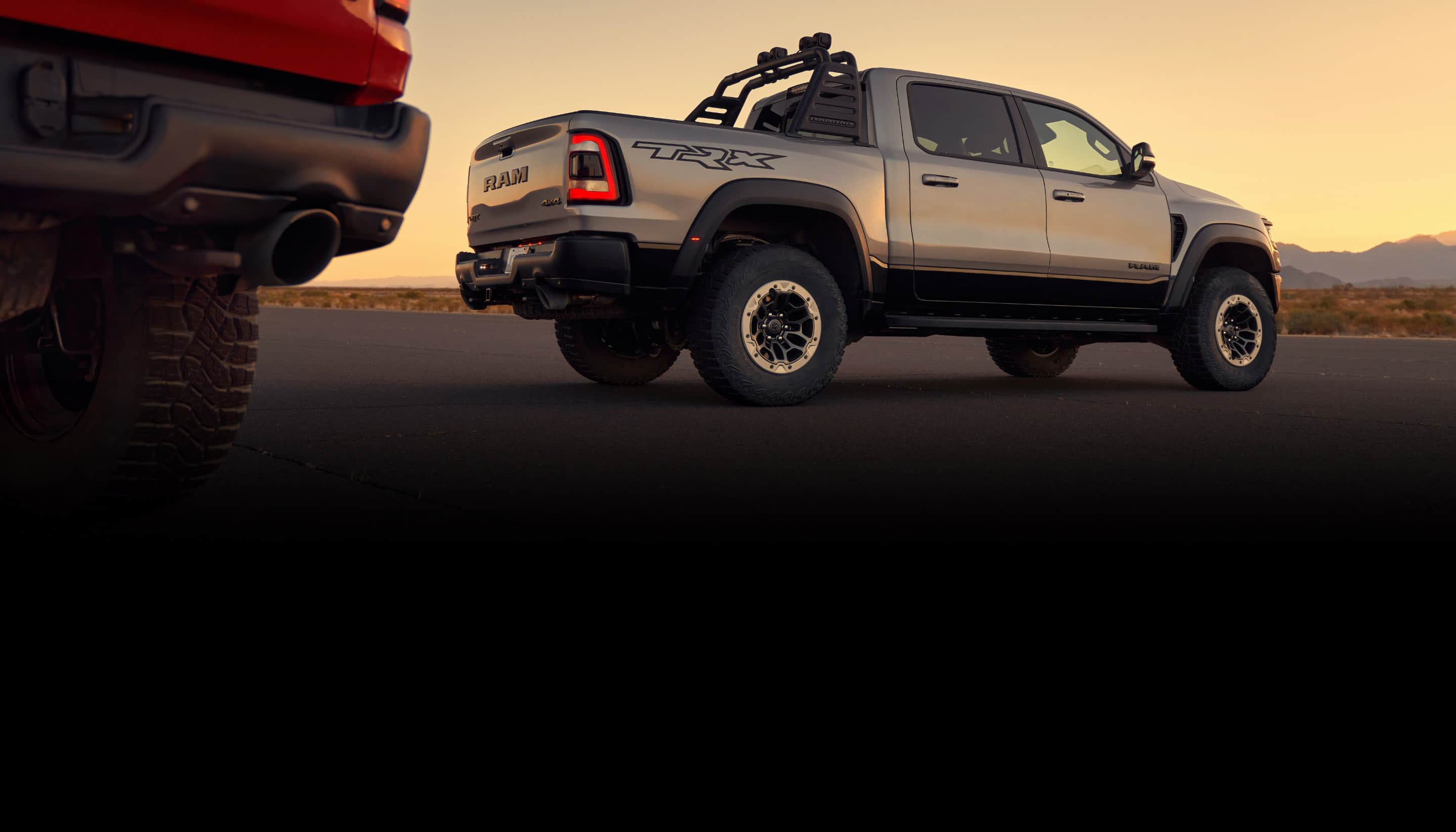 A silver 2021 Ram 1500 TRX with beadlock capable aluminum wheels and a Rambar by Mopar parked on blacktop at sunset, with the right rear corner of a red 2021 Ram 1500 TRX in the foreground.