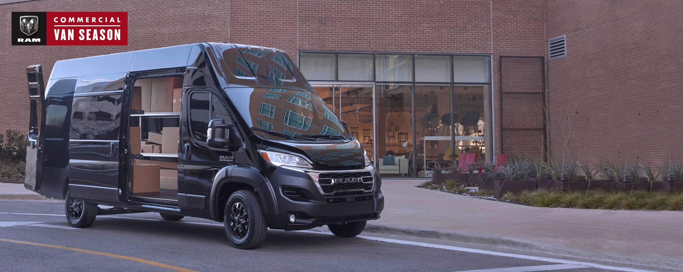 A 2023 Ram ProMaster 3500 Super High Roof Cargo Van parked next to a furniture store, its rear doors and passenger-side sliding door open, revealing shelves full of boxes. Ram Commercial Van Season.
