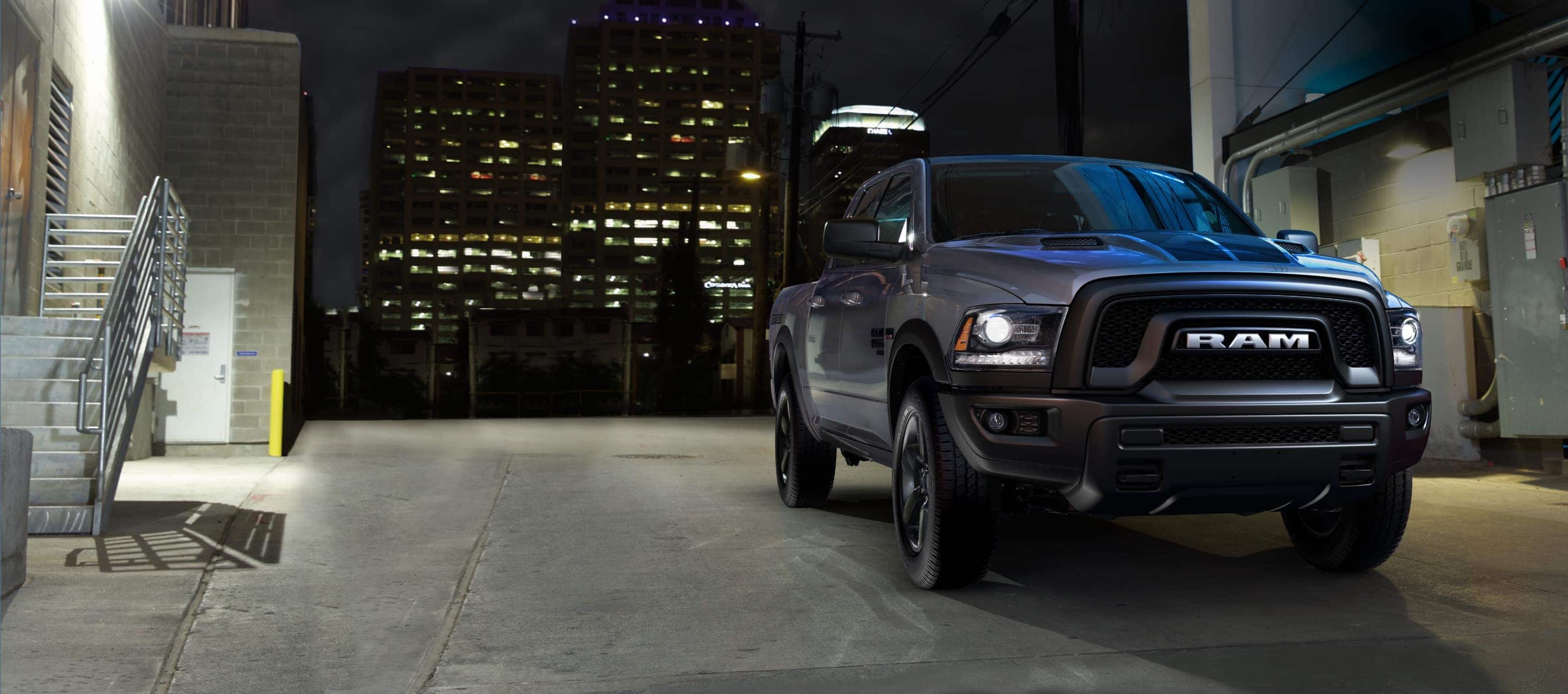  A black 2022 Ram 1500 Classic Warlock Crew Cab 4x4 with its headlamps on, being driven through an alleyway in an industrial area at night, with skyscrapers in the background. 