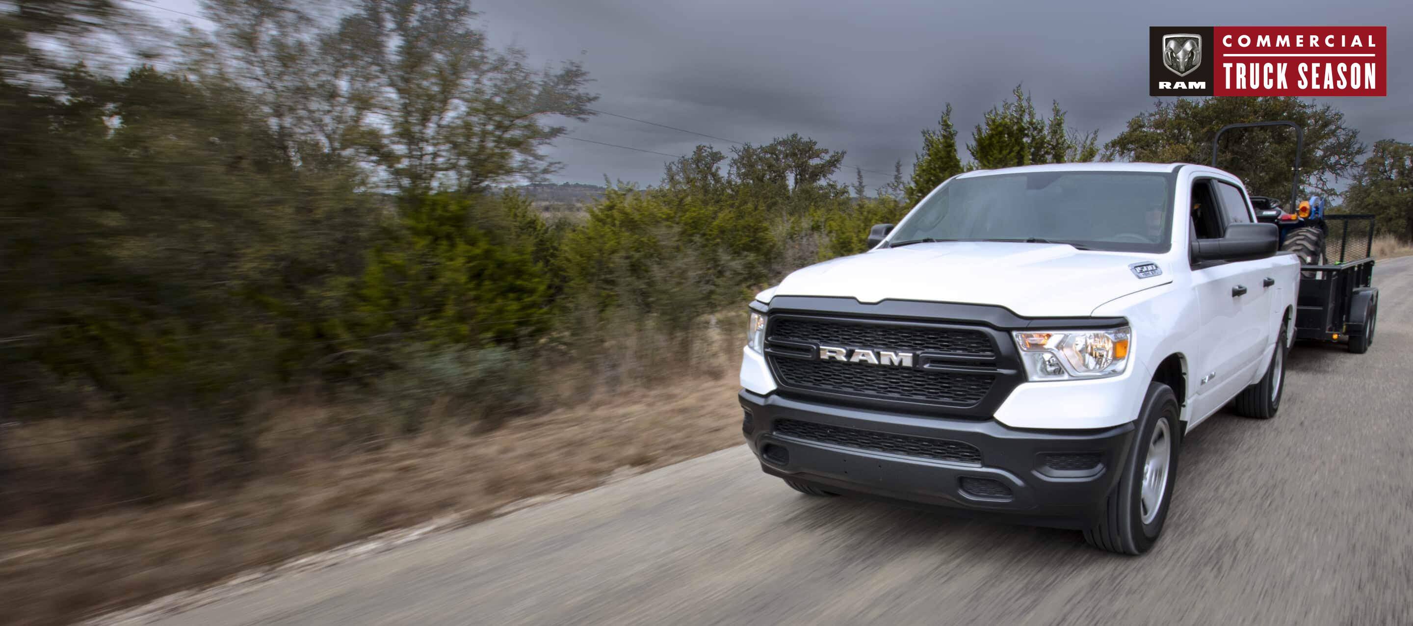 A white 2022 Ram 1500 Tradesman 4x4 Crew Cab towing an ATV on a flatbed trailer as it's driven on a country road with a stormy sky overhead. Ram Commercial Truck Season.