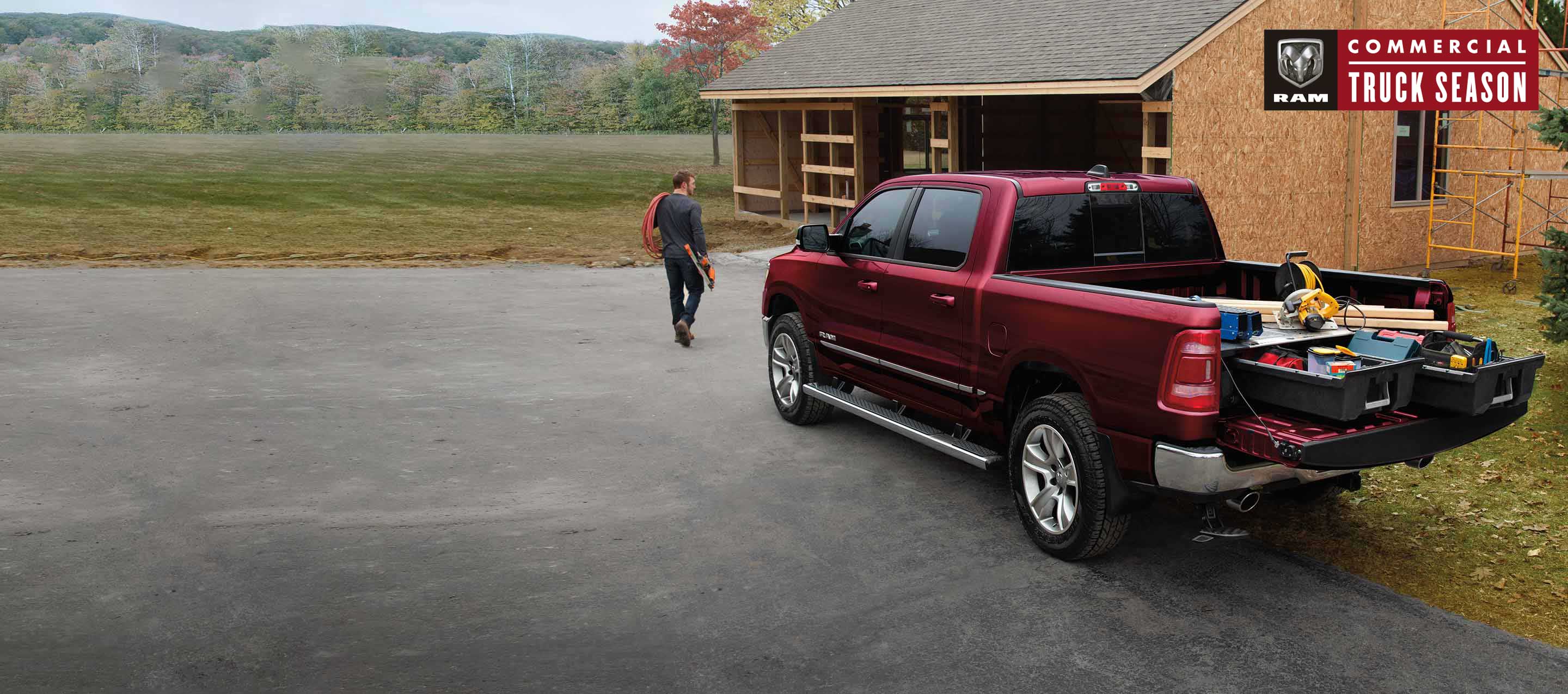 A red 2022 Ram 1500 Big Horn Crew Cab 4x4 with its tailgate open and its truck bed filled with tools, parked at a home under construction. Ram Commercial Truck Season.