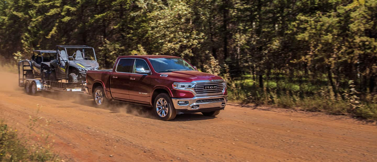 The 2023 Ram 1500 towing two ATVs on a flatbed as it's driven on a dirt road.