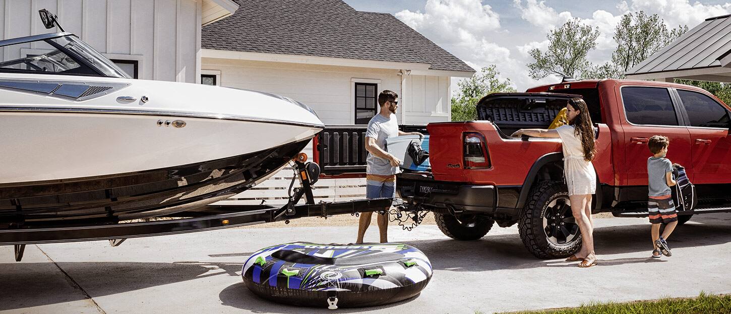 A man loading a wakeboard through the open tailgate door of a 2022 Ram 1500 with an attached boat trailer.