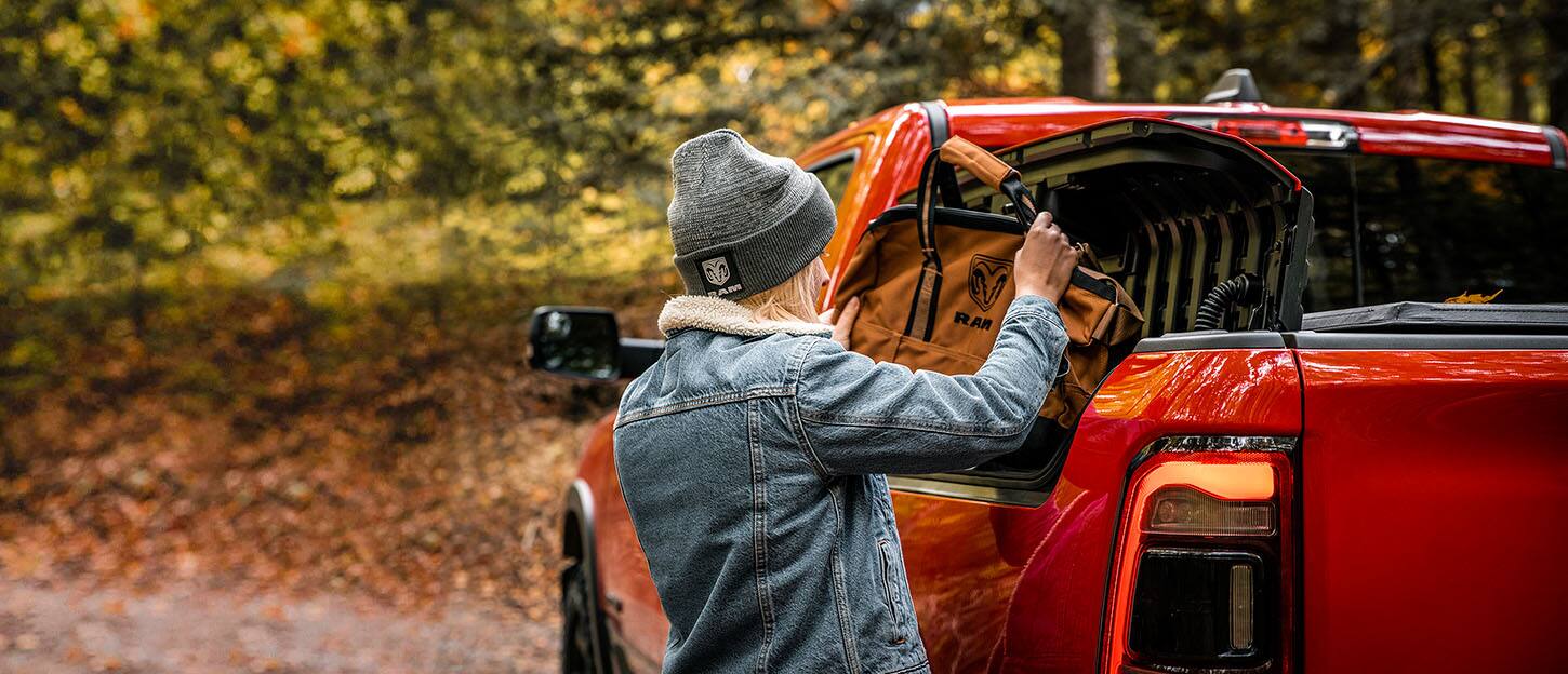A woman loading a canvas tool bag into the open RamBox cargo compartment on the driver's side of the 2022 Ram 1500 pickup bed.