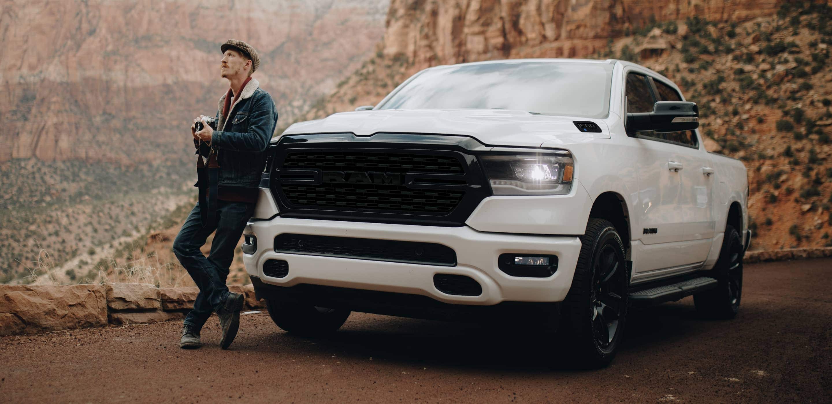 Display A man with a camera leaning against a 2022 Ram 1500, looking up at the mountains surrounding him.