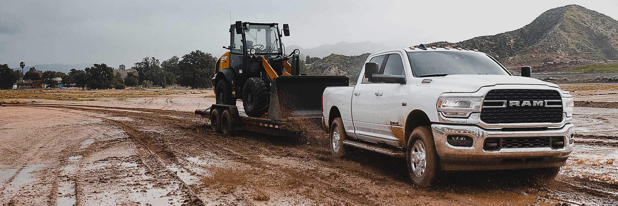 The 2022 Ram 2500 towing a flatbed with a digger on it through a muddy field.
