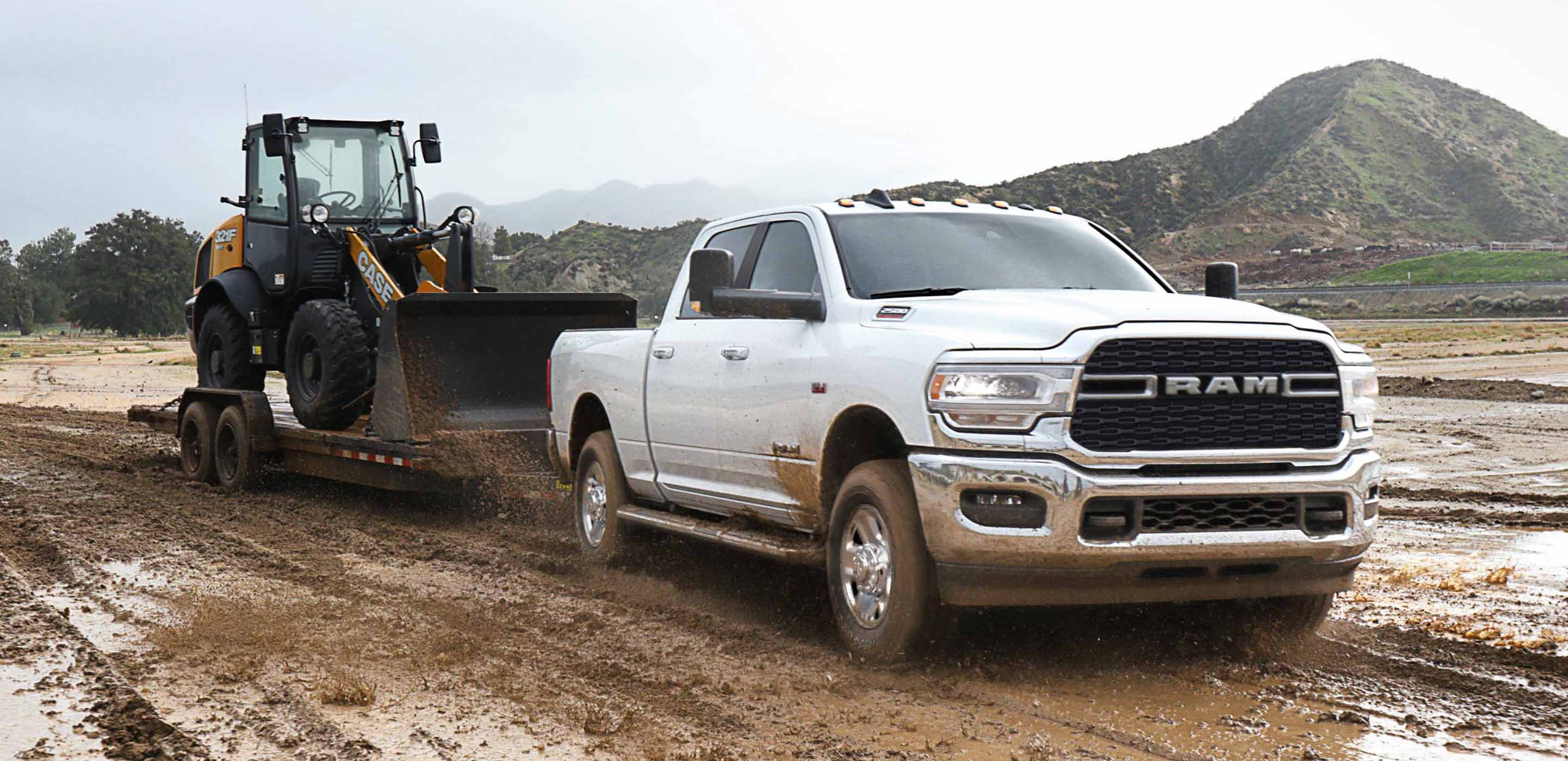 Display The 2022 Ram 2500 towing a flatbed trailer with an excavator on it through a muddy field.