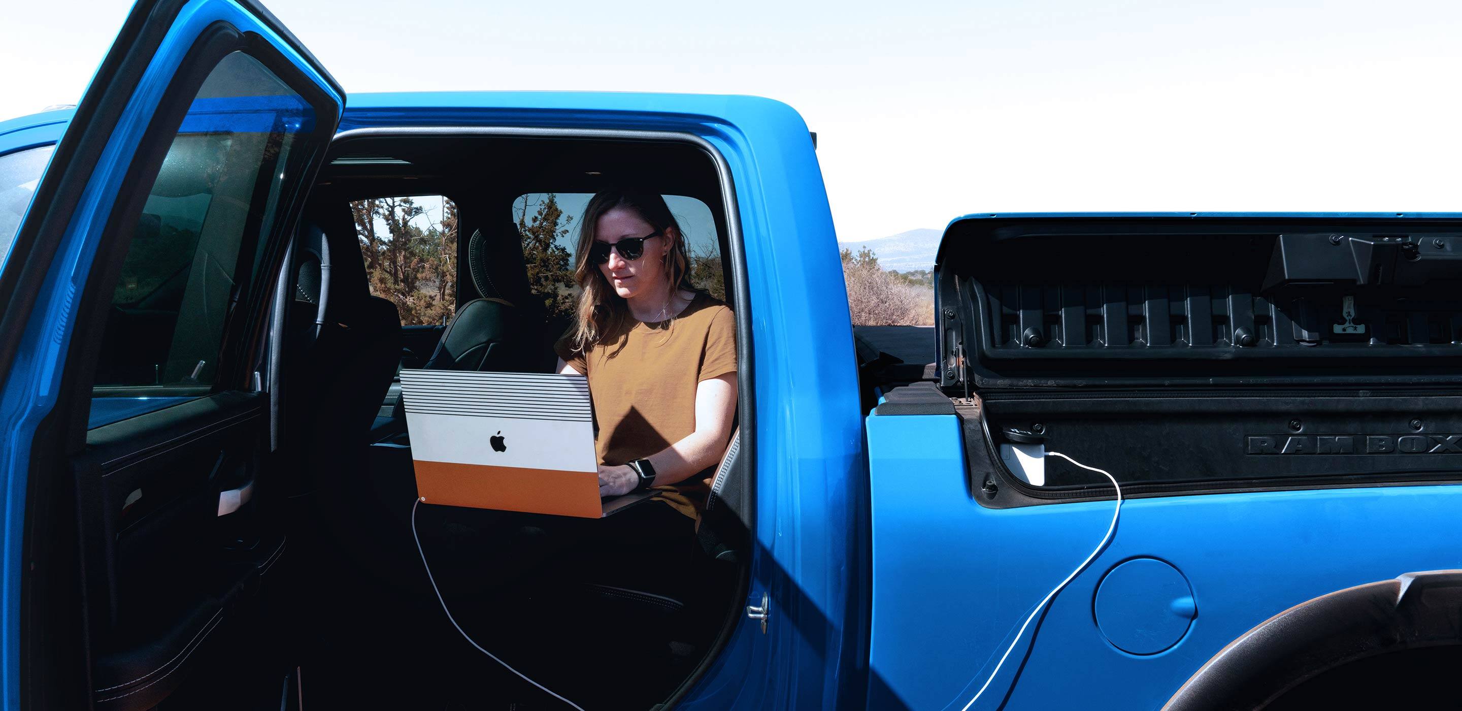 Display A woman in a parked 2022 Ram 2500, working on a laptop that is plugged into the outlet in the Rambox Cargo Management System.