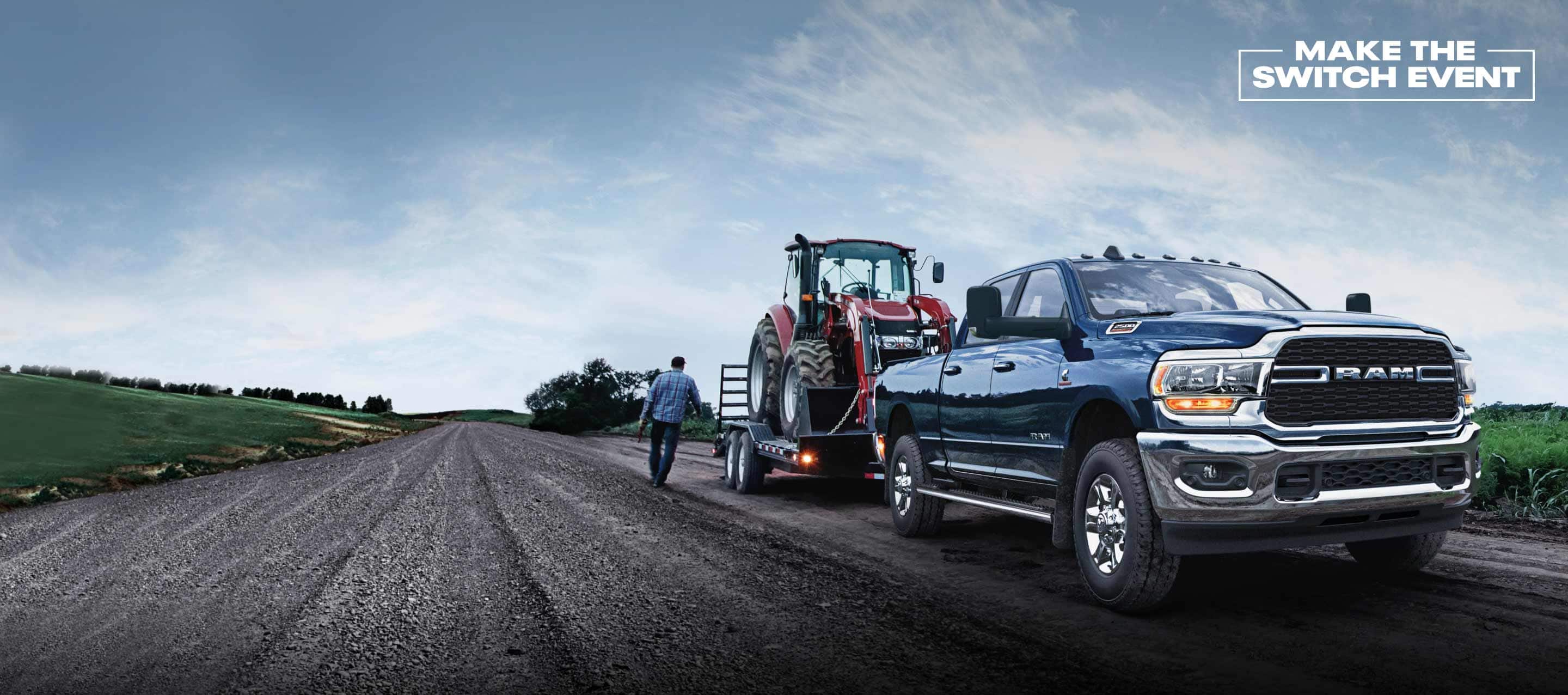 The 2022 Ram 2500 towing a flatbed with a tractor on it. Make the Switch Event logo.