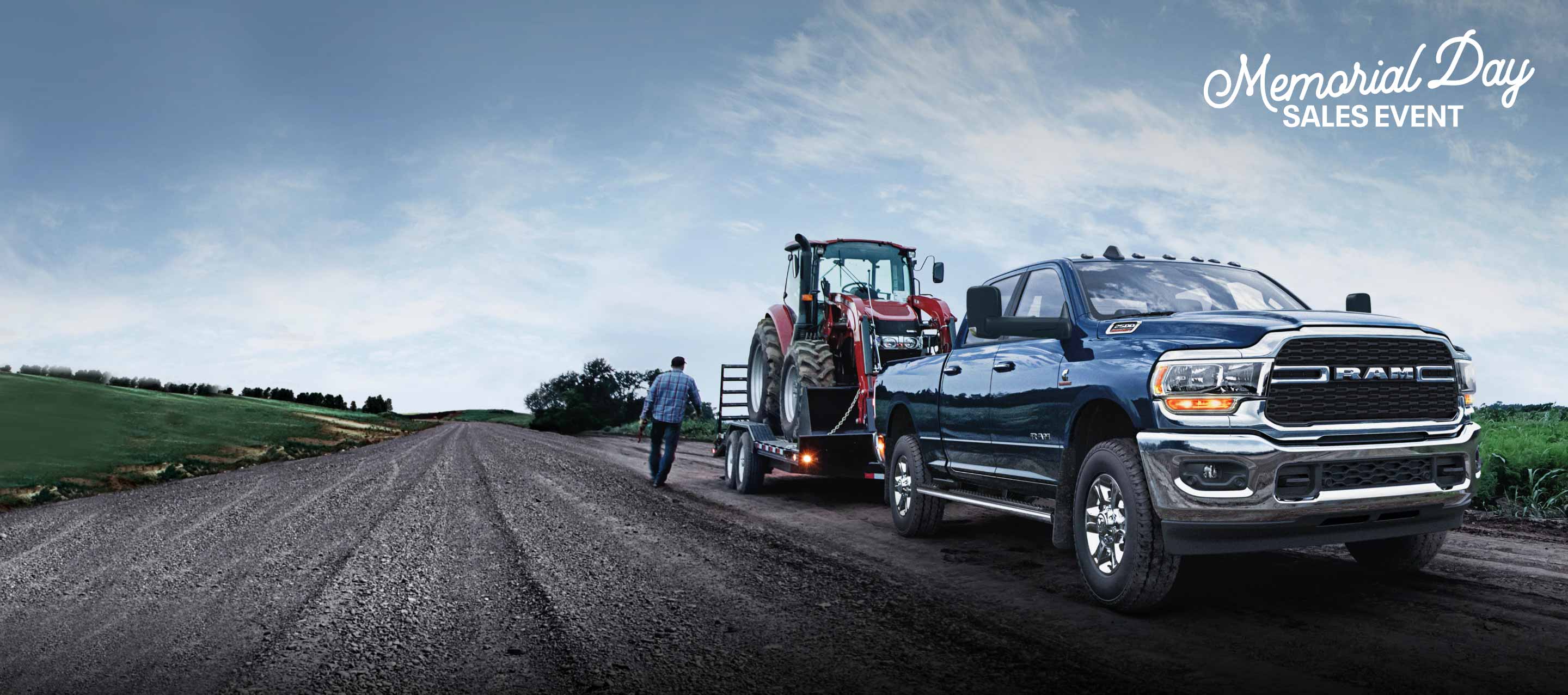 The 2022 Ram 2500 towing a flatbed with a tractor on it. Memorial Day Sales Event logo.