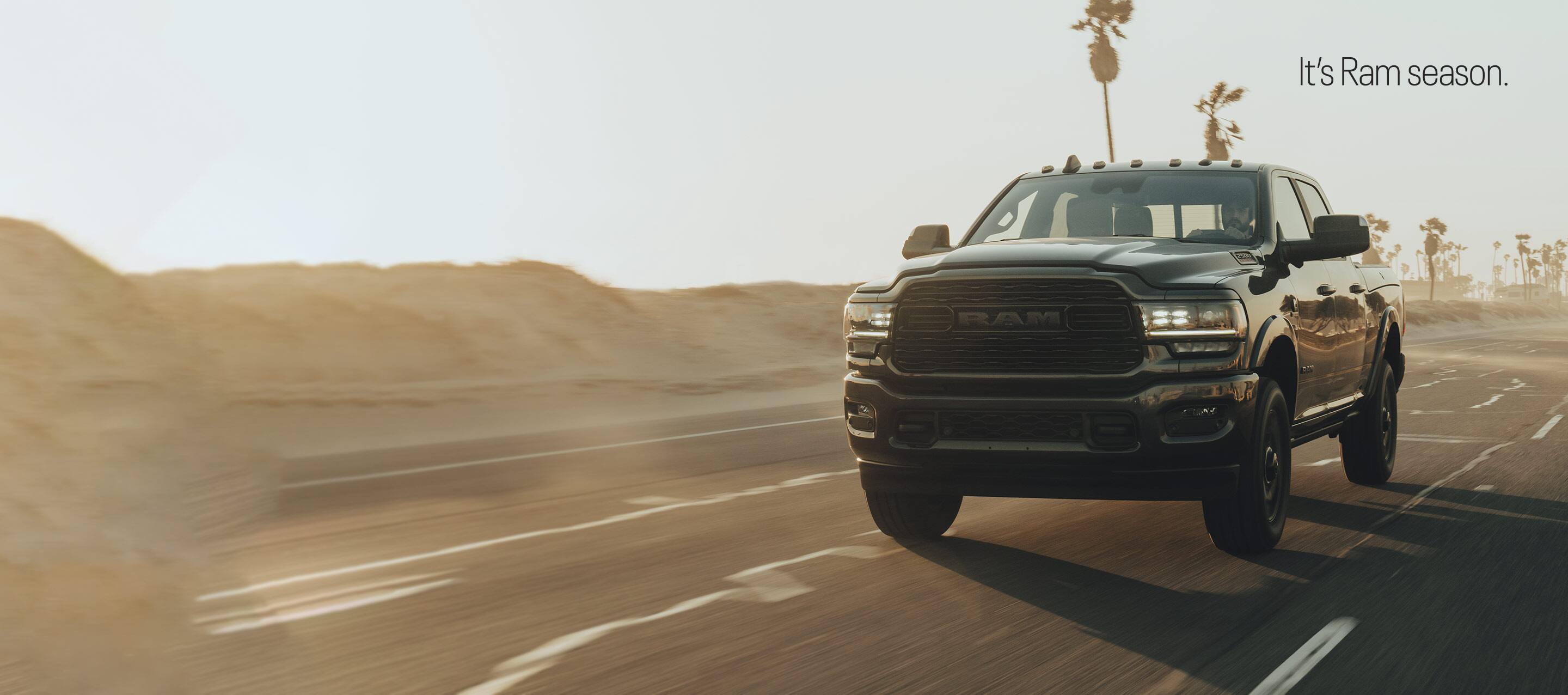 A 2022 Ram 2500 Limited 4x4 Crew Cab being driven on a highway with sand hills and palm trees in the background. It's Ram Season.