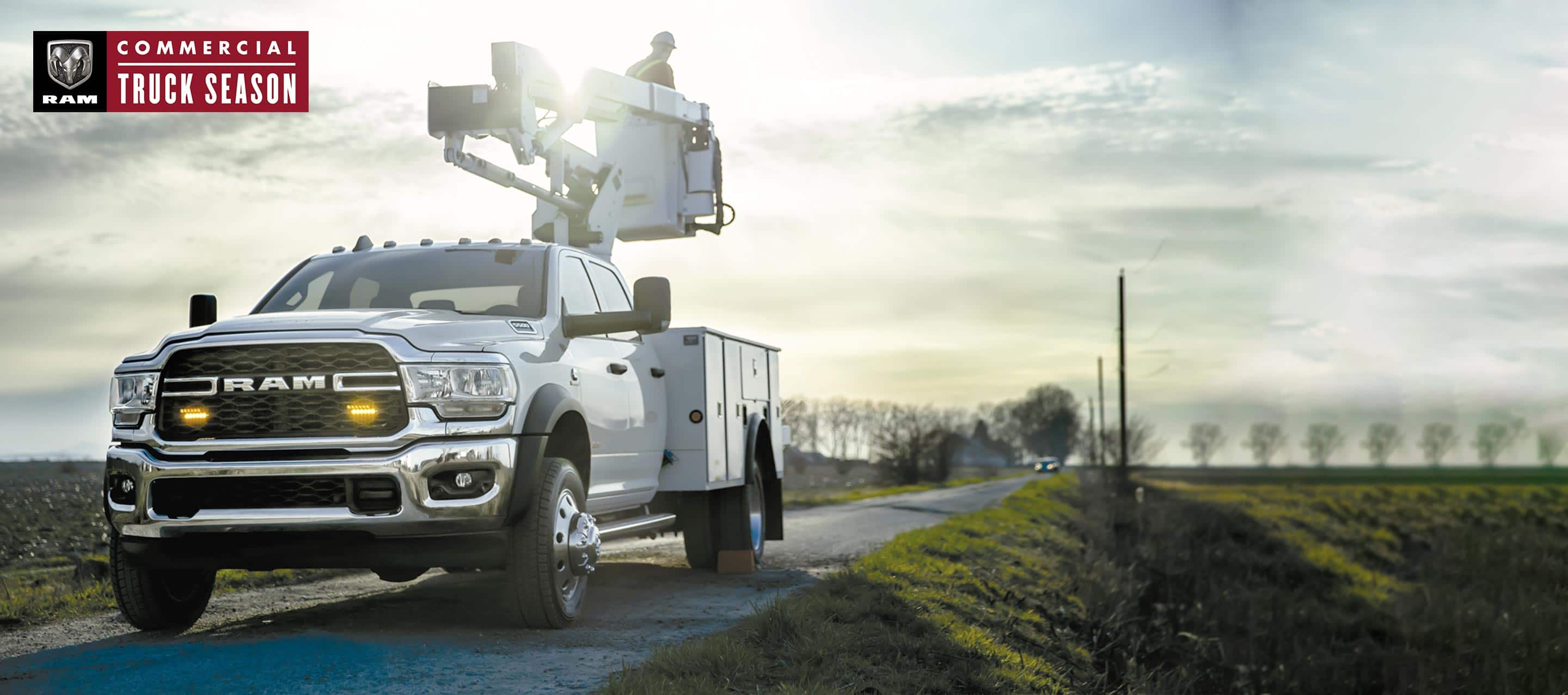 A white 2022 Ram 5500 Tradesman Chassis Cab 4x4 Crew Cab with mechanical upfit, parked on a single lane road, with a technician standing in the cherry picker bucket. Ram Commercial Truck Season.