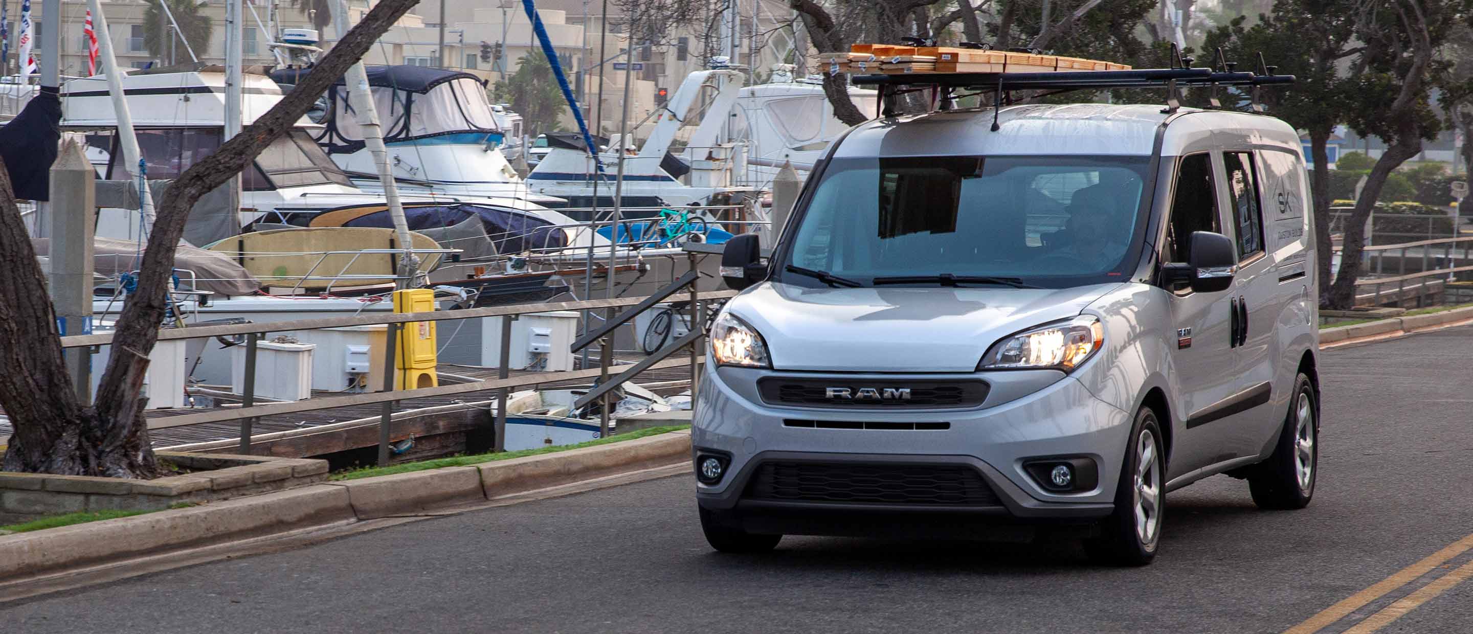 A 2022 Ram ProMaster City, carrying planks of lumber on its roof rack as it's driven past a marina.