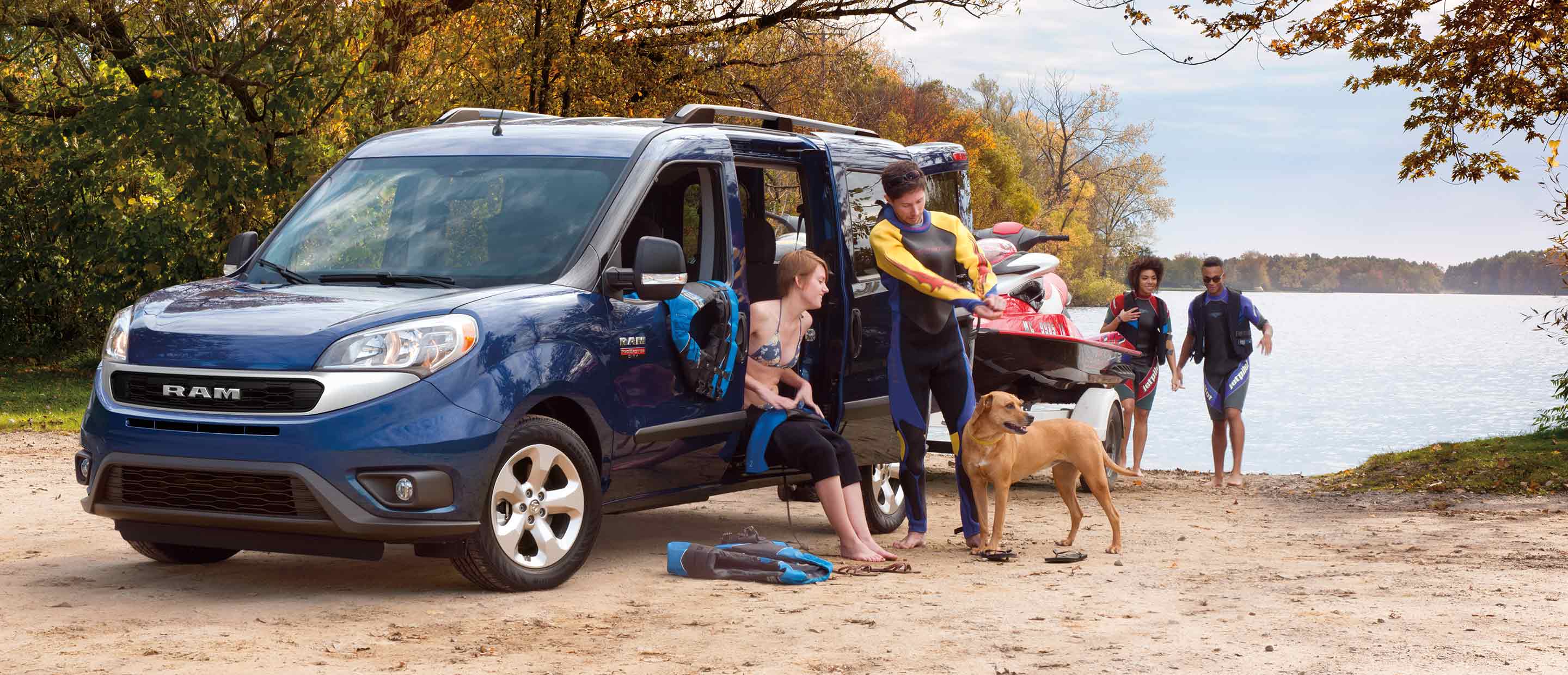 A 2022 Ram ProMaster City Wagon parked on a beach with a jet ski on a trailer behind it and a man and woman putting on wetsuits beside the vehicle.