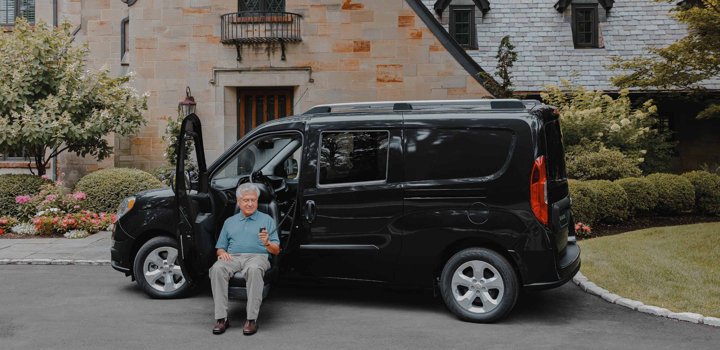 Display The 2022 Ram ProMaster City with a man sitting in a wheelchair transfer seat, holding the controls to lift his seat into the driver's position.