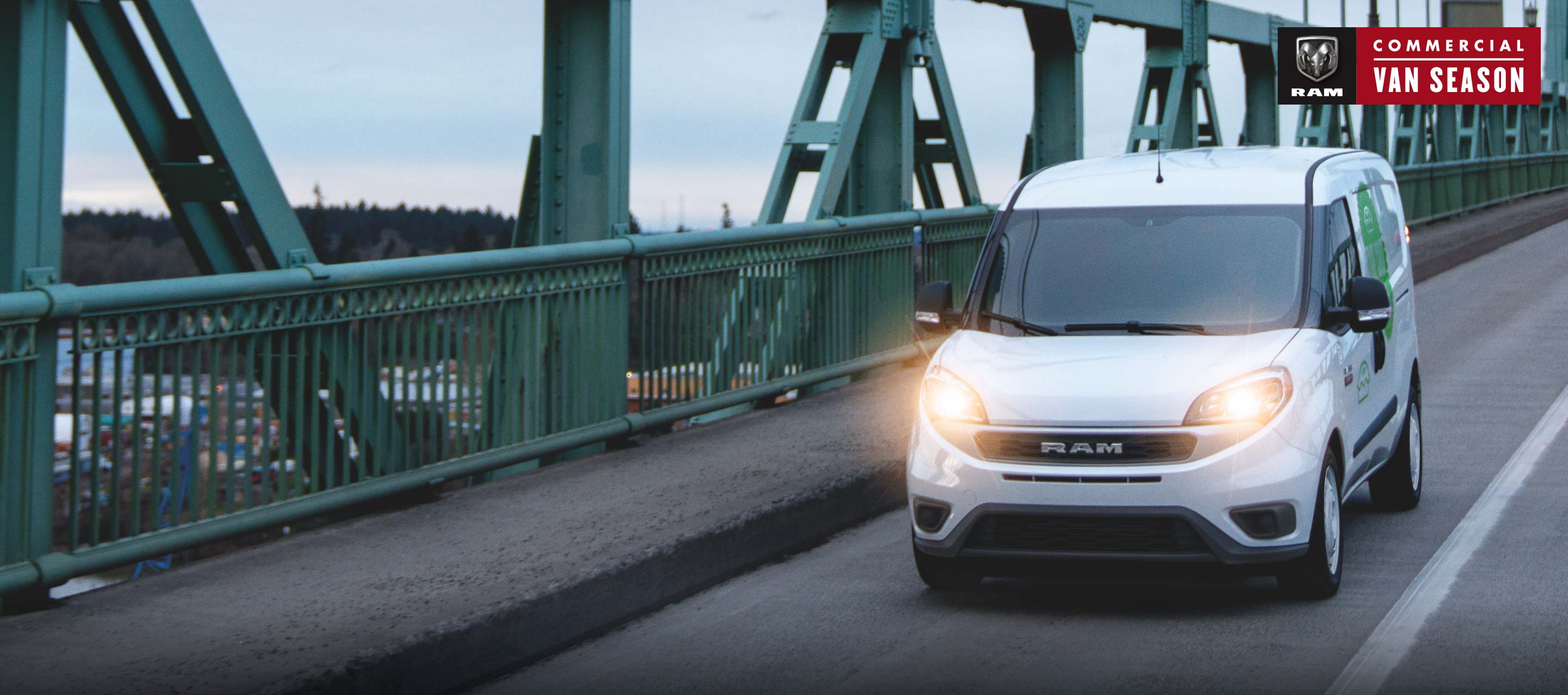 Ram Commercial Van season. A white 2022 Ram ProMaster City Cargo Van being driven across a bridge with its headlamps on.