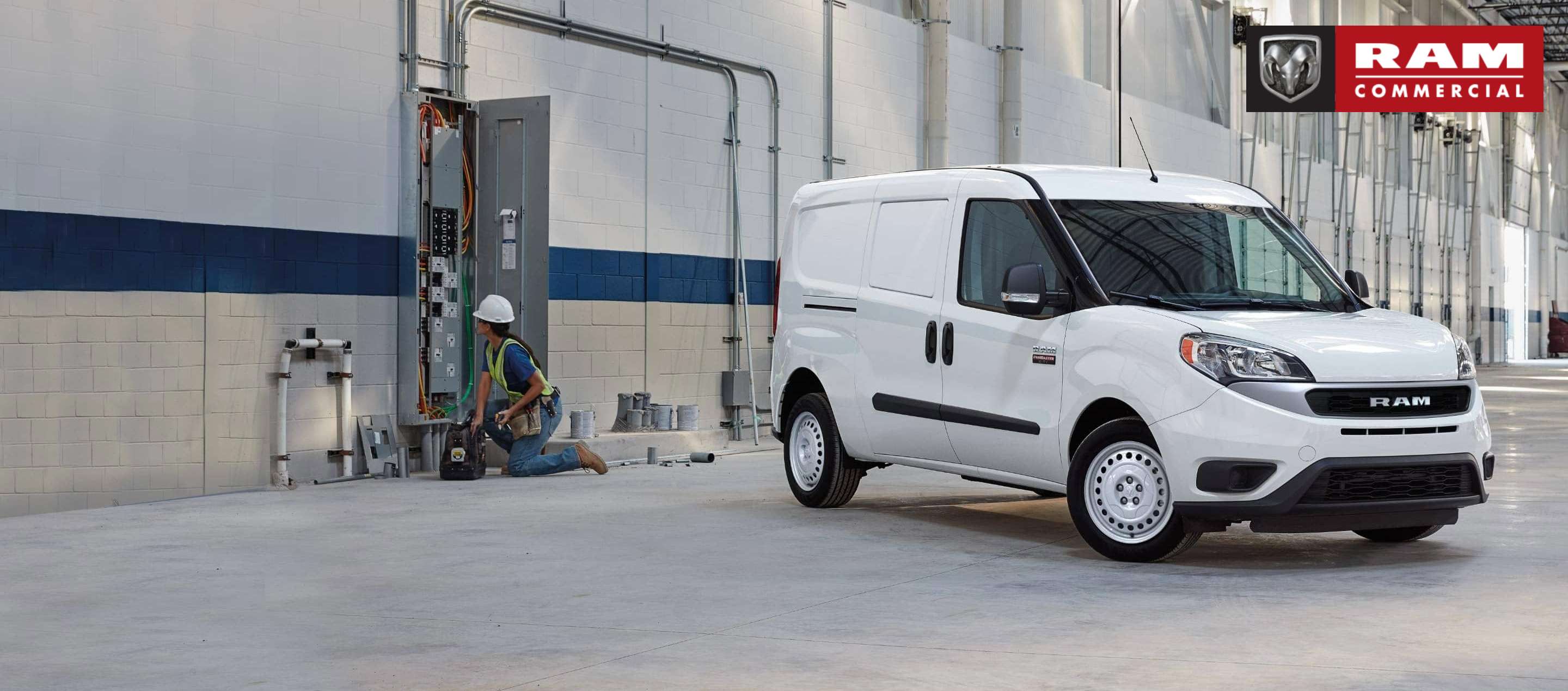 A white 2022 Ram ProMaster City Cargo Van in a large industrial garage with a technician nearby, working on an electrical breaker box. Ram Commercial.