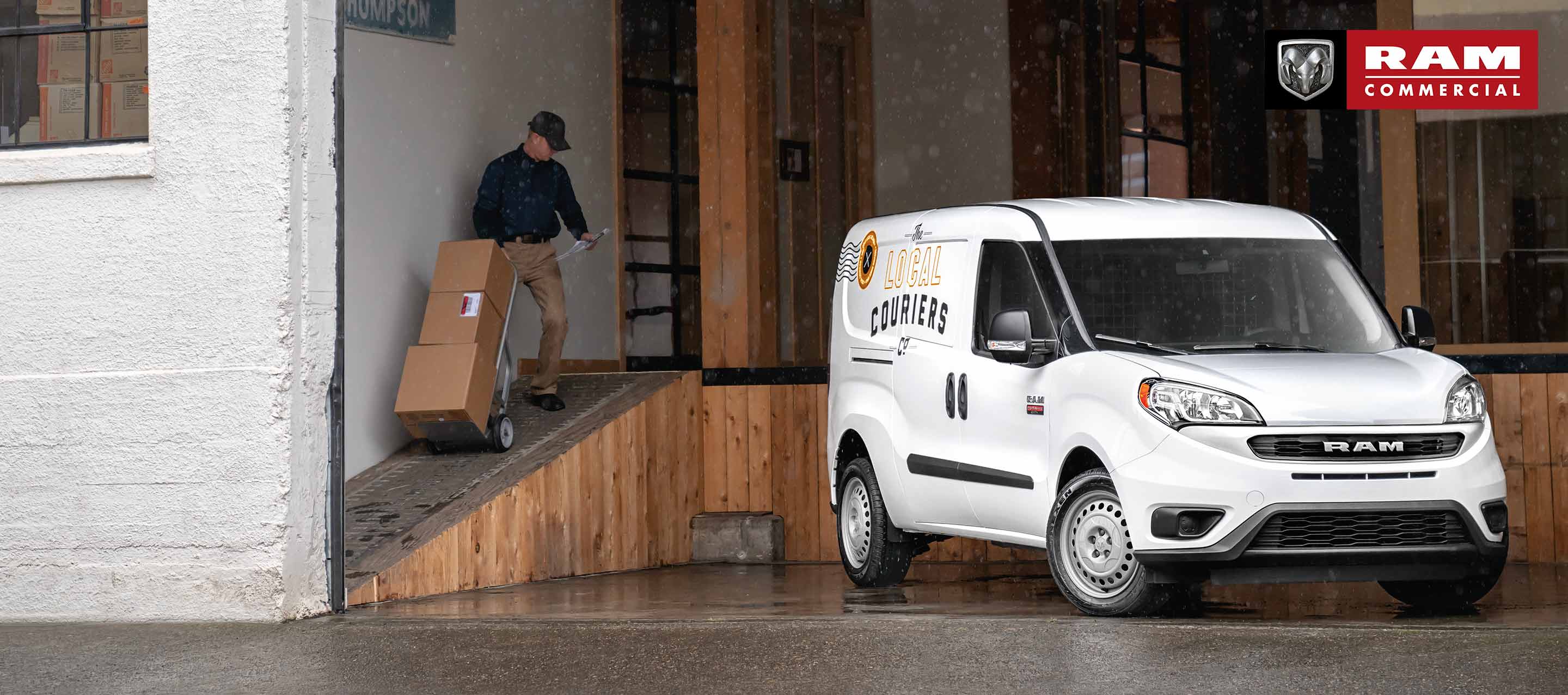 A white 2022 Ram ProMaster City Cargo Van with delivery company signage on its side, parked at a loading dock as a delivery man hauls a set of boxes up a ramp. Ram Commercial.