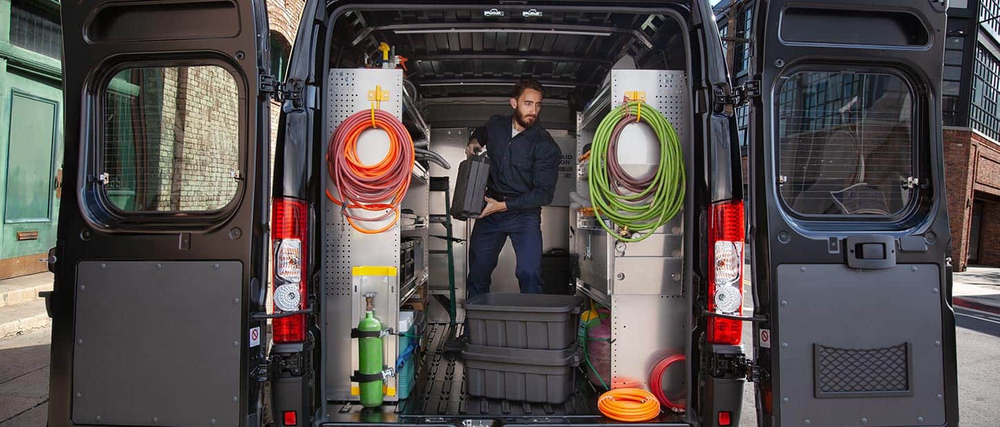 A 2022 Ram ProMaster with its cargo compartment upfitted with shelving and hooks for cables. A man stands upright in the van.