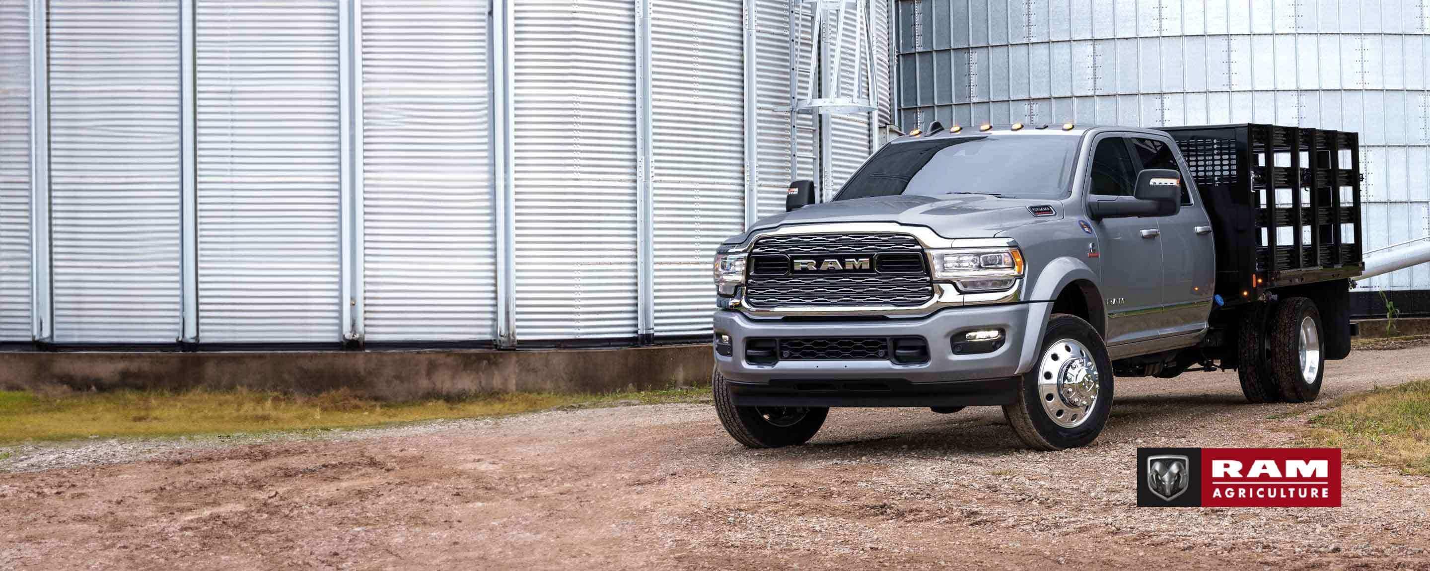 A silver 2024 Ram Limited Crew Cab Chassis Cab with a utility trailer, parked beside a farm's silo. Ram Agriculture.