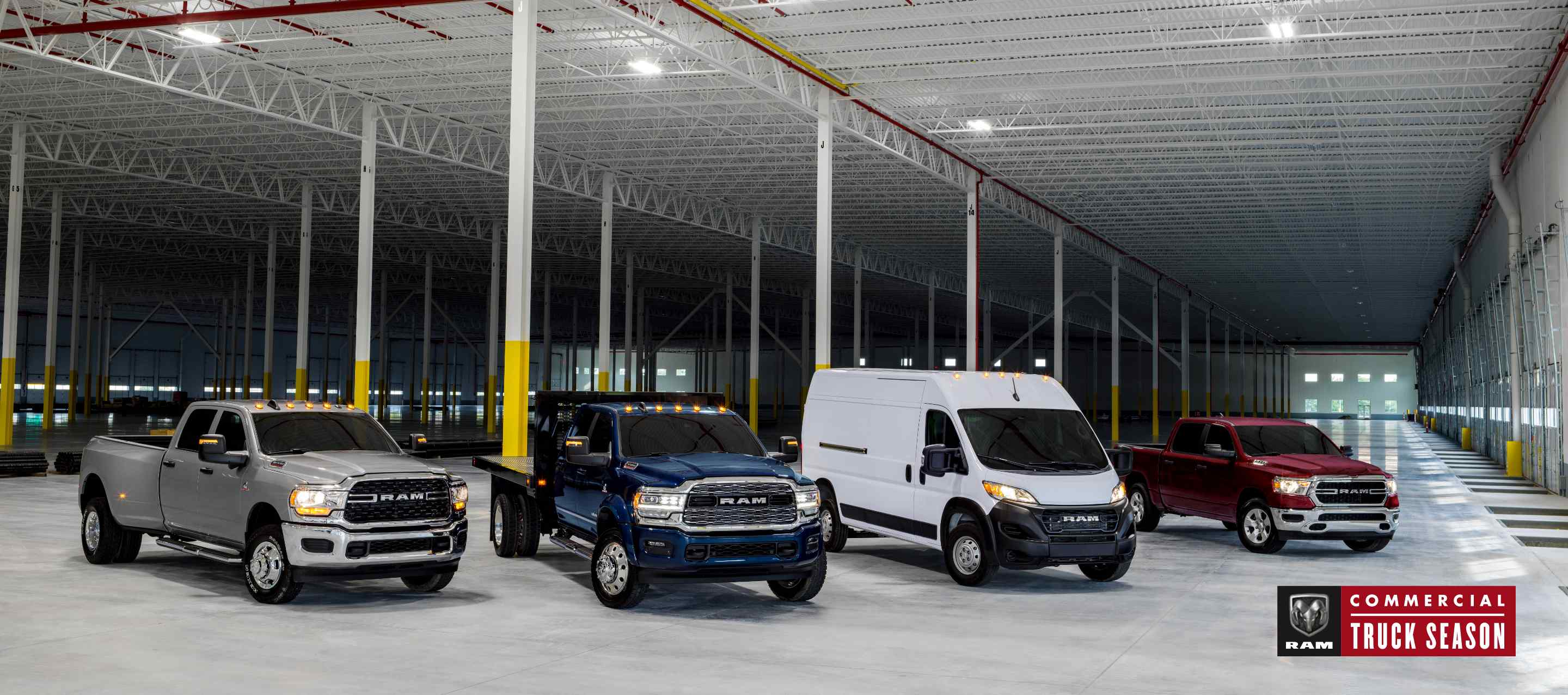 A lineup of four Ram Brand vehicles parked in an industrial garage. From left to right: a silver 2023 Ram 3500 Big Horn 4x4 Crew Cab, a blue 2023 Ram 5500 Limited 4x4 Chassis Crew Cab with utility bed upfit, a white 2023 Ram ProMaster 2500 Cargo Van High Roof and a red 2023 Ram 1500 Tradesman 4x4 Crew Cab. Ram Commercial Truck Season.