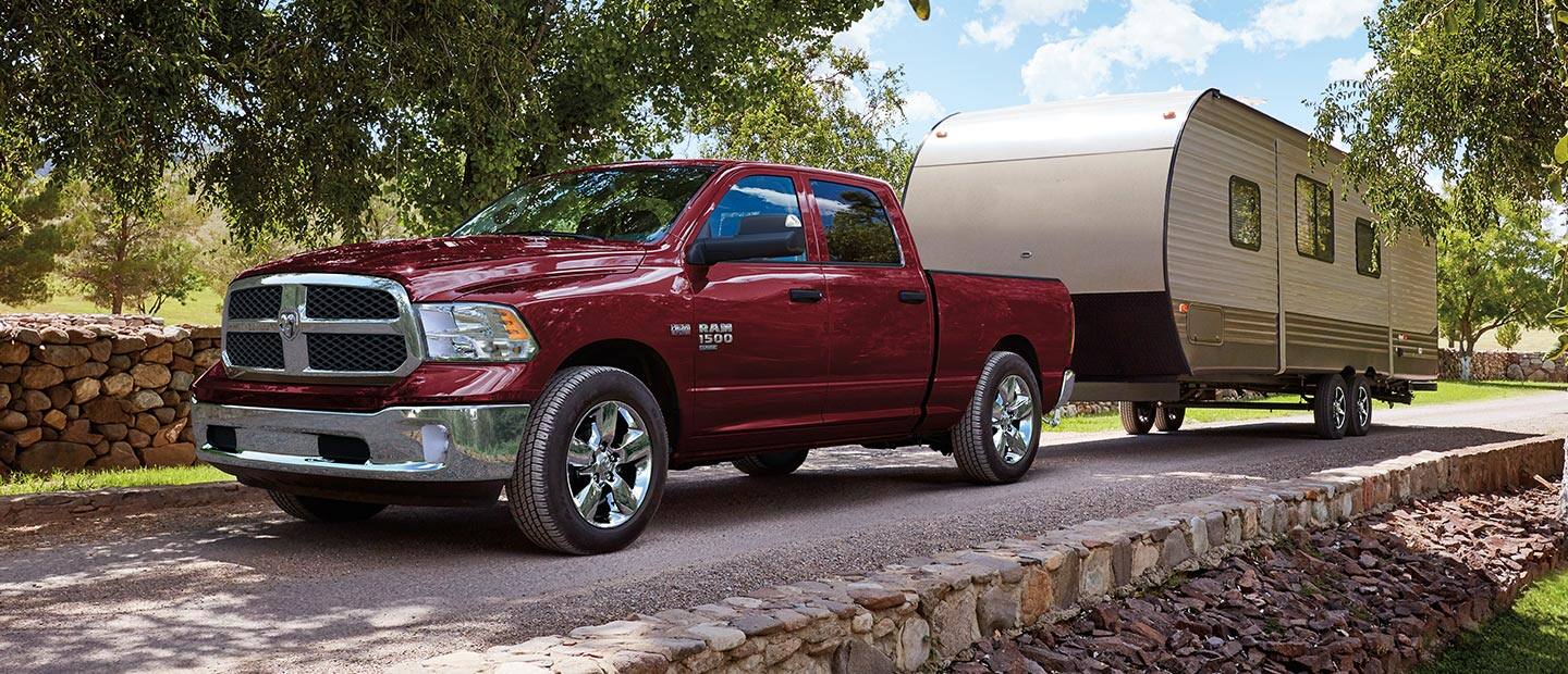 A red 2023 Ram 1500 Classic TBD Crew Cab being driven down a road beside a cobblestone fence,  towing a travel trailer.