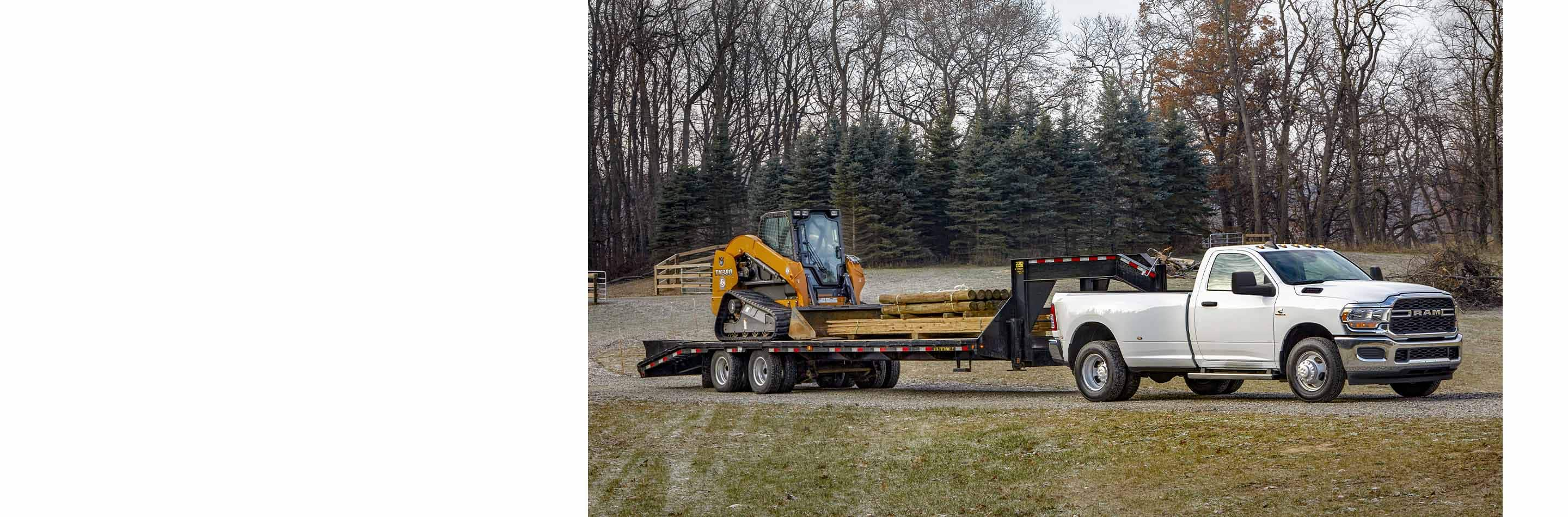 A profile of a 2023 Ram 3500 Big Horn Regular Cab being driven down a highway, towing a fifth wheel extended flatbed trailer with an excavator on it. 