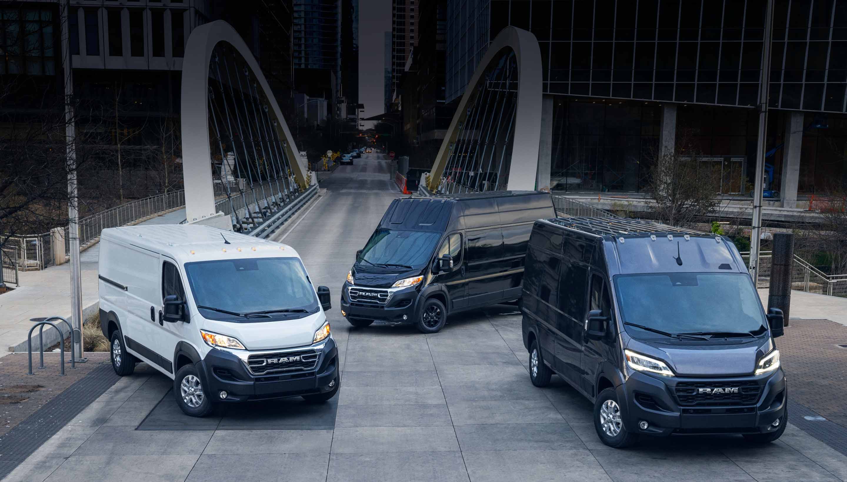 Three 2023 Ram ProMaster models: the standard roof, the high roof and the super high roof parked just beyond a bridge in the center of a busy large city.