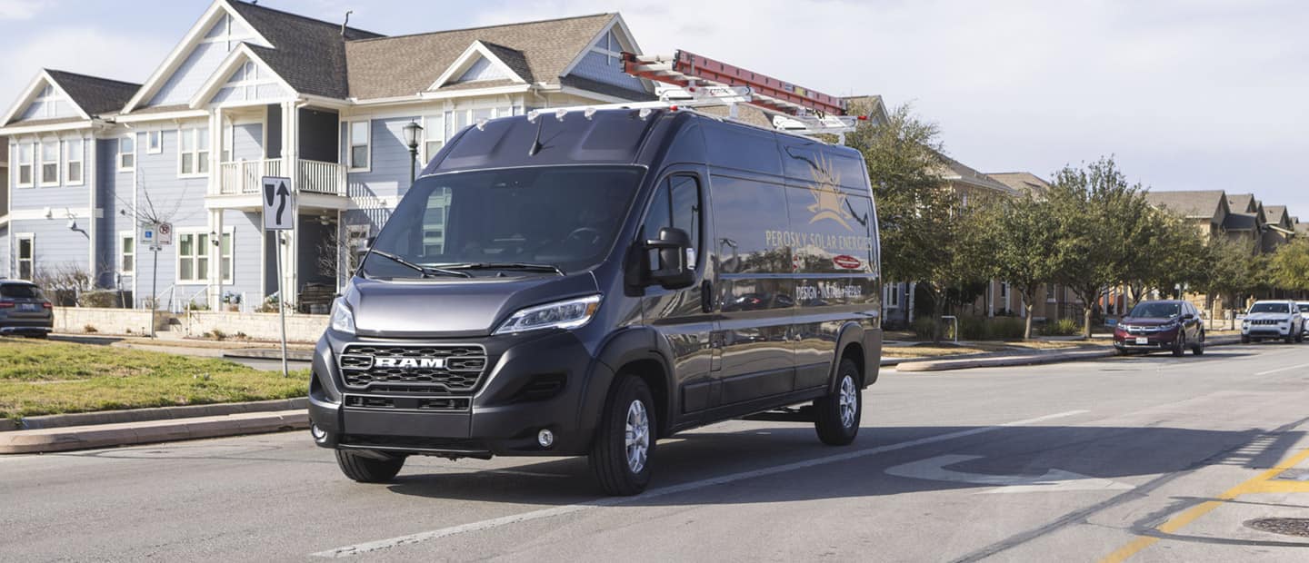 The 2023 Ram ProMaster High Roof with a ladder attached to its roof rack being driven through a neighborhood.