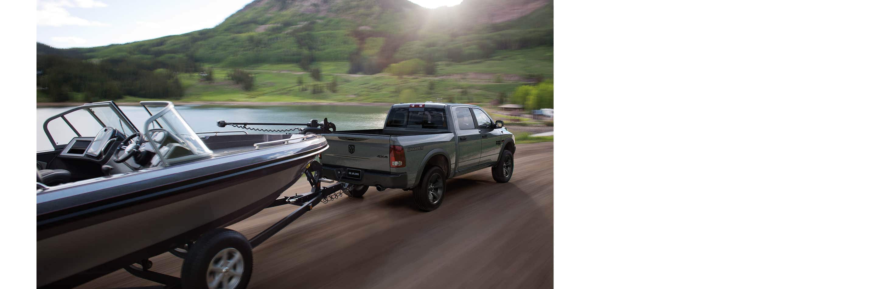 Rear view of a 2023 Ram 1500 Classic Warlock 4x4 Crew Cab being driven beside a lake, towing a motorboat.