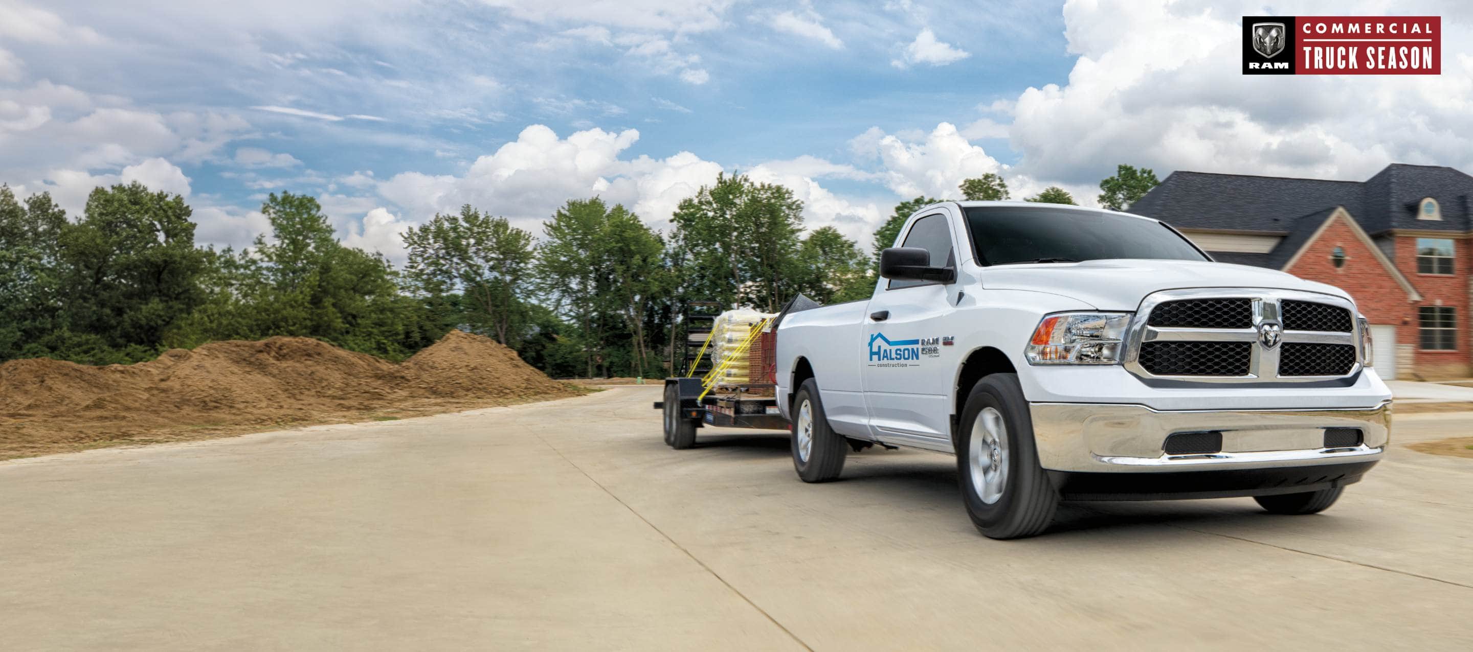 A white 2023 Ram 1500 Classic Tradesman 4x4 Regular Cab with a construction company's signage on its driver's door, towing a flatbed trailer carrying construction supplies as it pulls away from a residential neighborhood. Ram Commercial Truck Season.