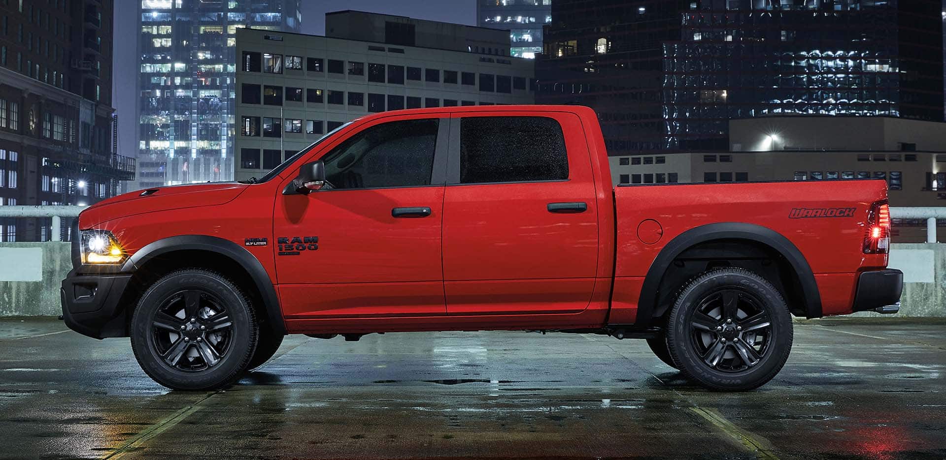 Display A profile of a red 2023 Ram 1500 Classic Warlock Crew Cab parked on a rooftop garage at night with high-rise buildings in the background.