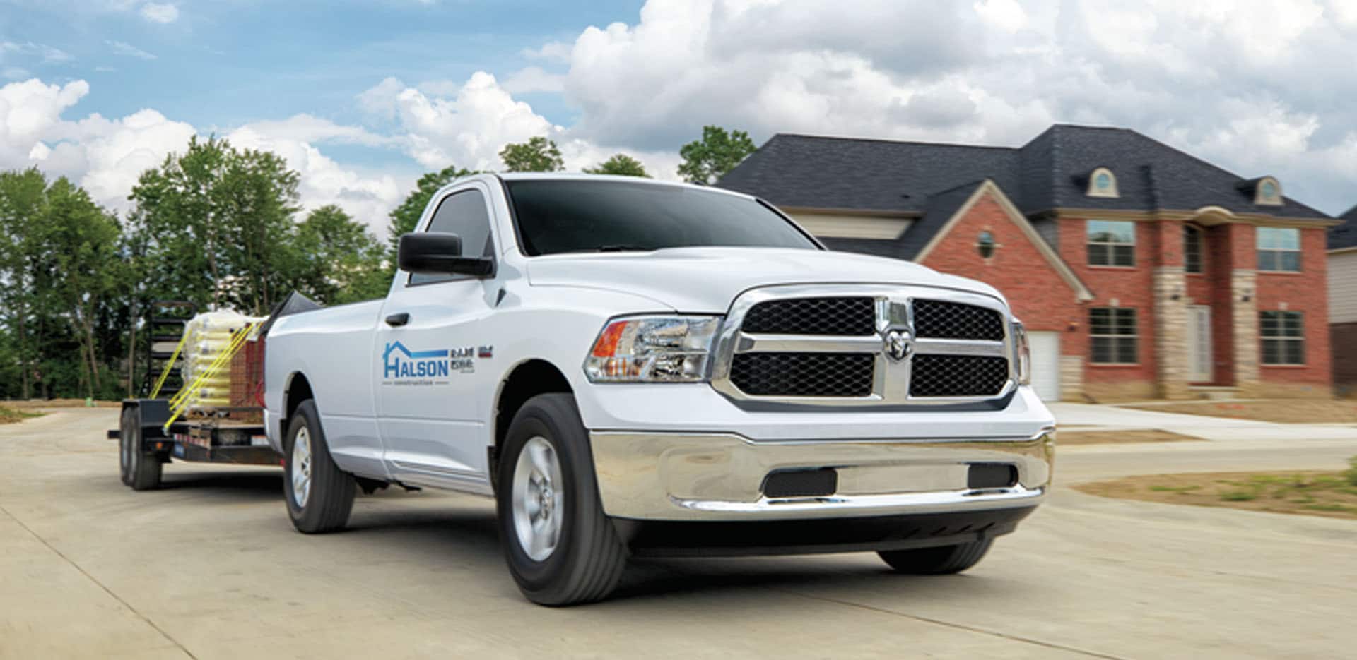 Display A white 2023 Ram 1500 Classic Tradesman Regular Cab with construction company signage on the passenger door, towing building materials as it is driven through a residential neighborhood.