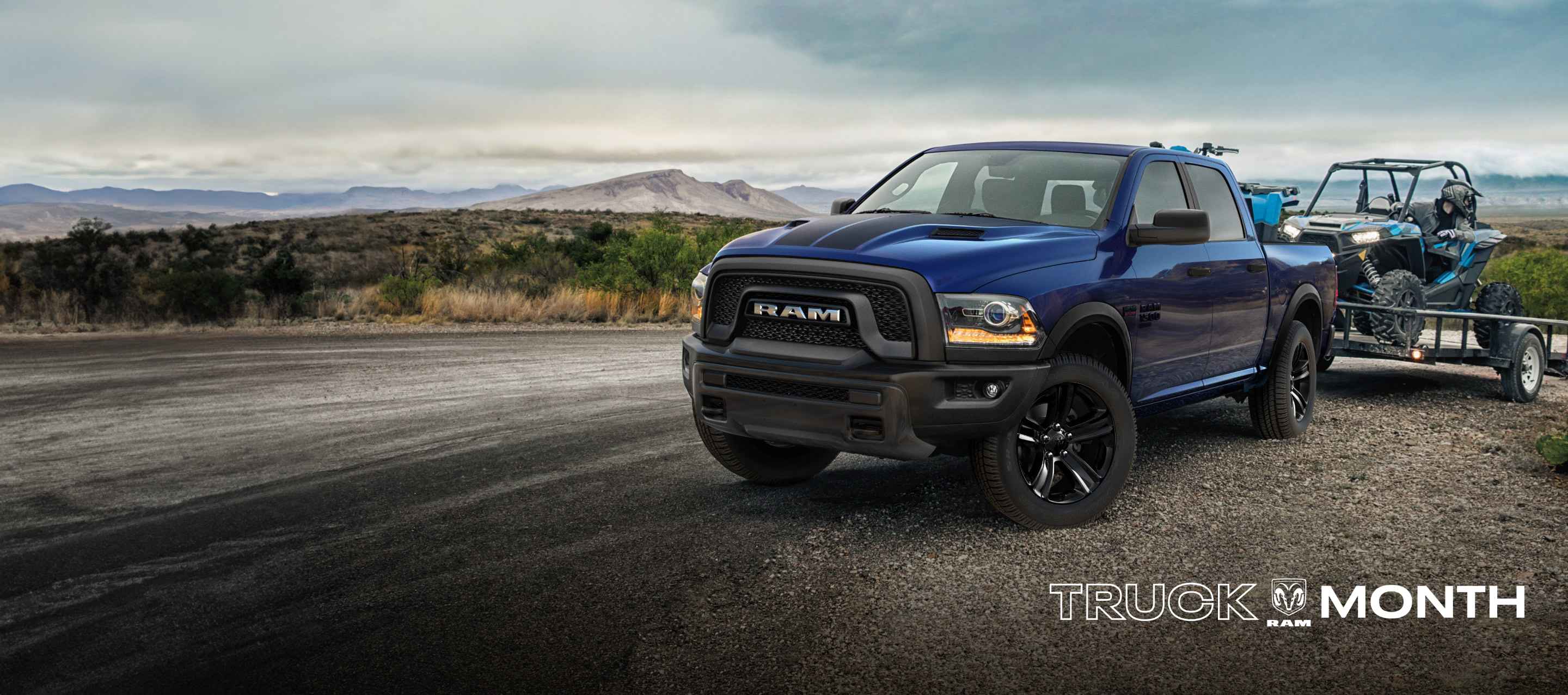A blue 2023 Ram 1500 Classic Warlock 4x4 Crew Cab parked on the shoulder of a highway, towing an ATV on a utility trailer.  Truck Month Sales Event.