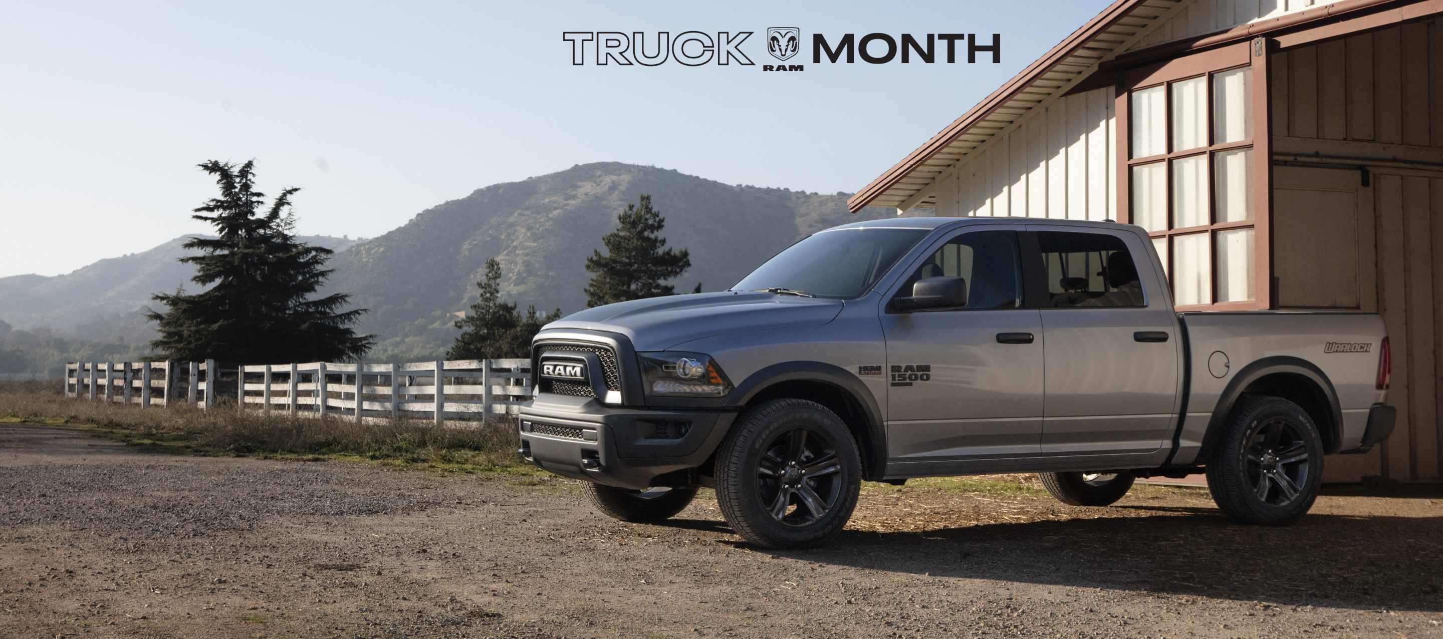 A driver-side profile of a silver 2023 Ram 1500 Classic Warlock Crew Cab parked on a farm, outside of a stable, with mountains in the background. Truck Month Sales Event.