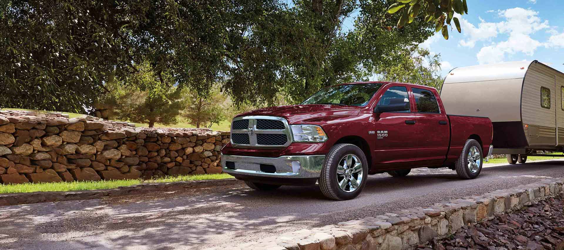 A red 2023 Ram 1500 Classic Tradesman 4x4 Crew Cab being driven down a road beside a cobblestone fence,  towing a travel trailer.