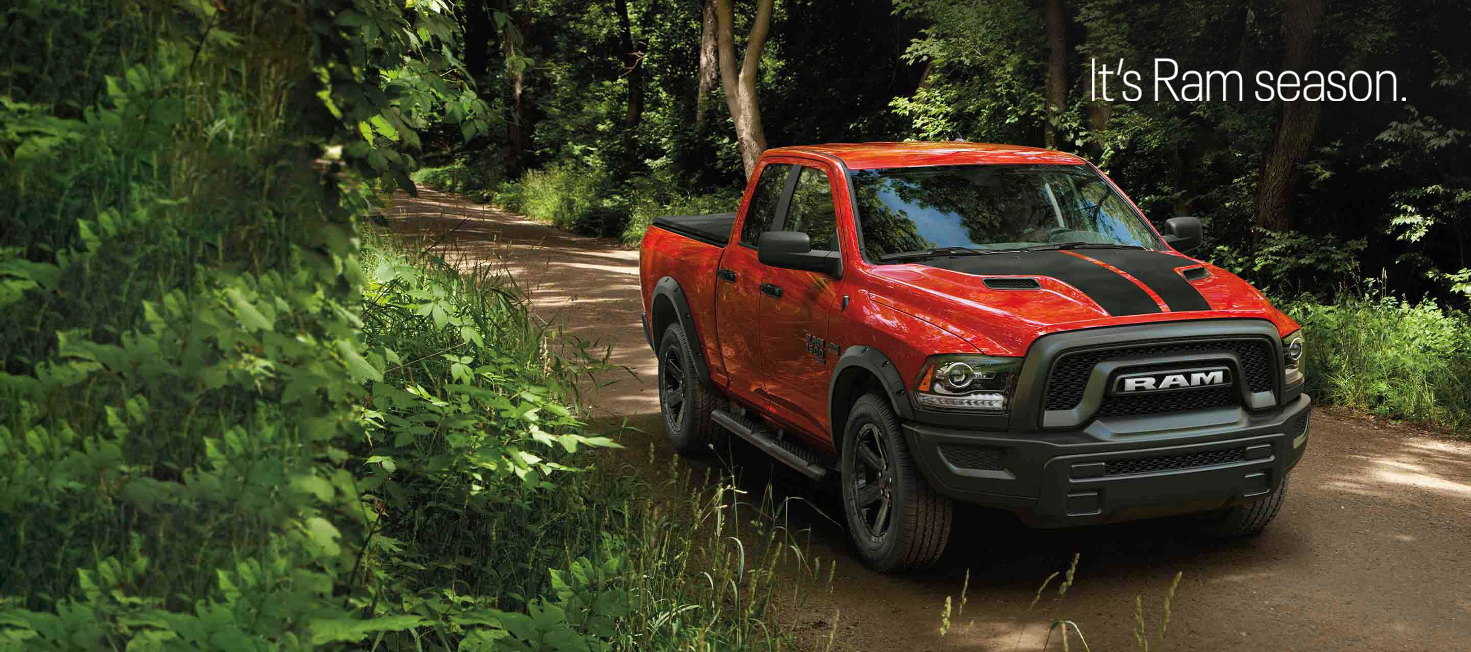 A red 2023 Ram 1500 Classic Warlock 4x4 Quad Cab with dual black center hood stripes and Mopar accessories, being driven through the woods on a dirt road. It's Ram season.