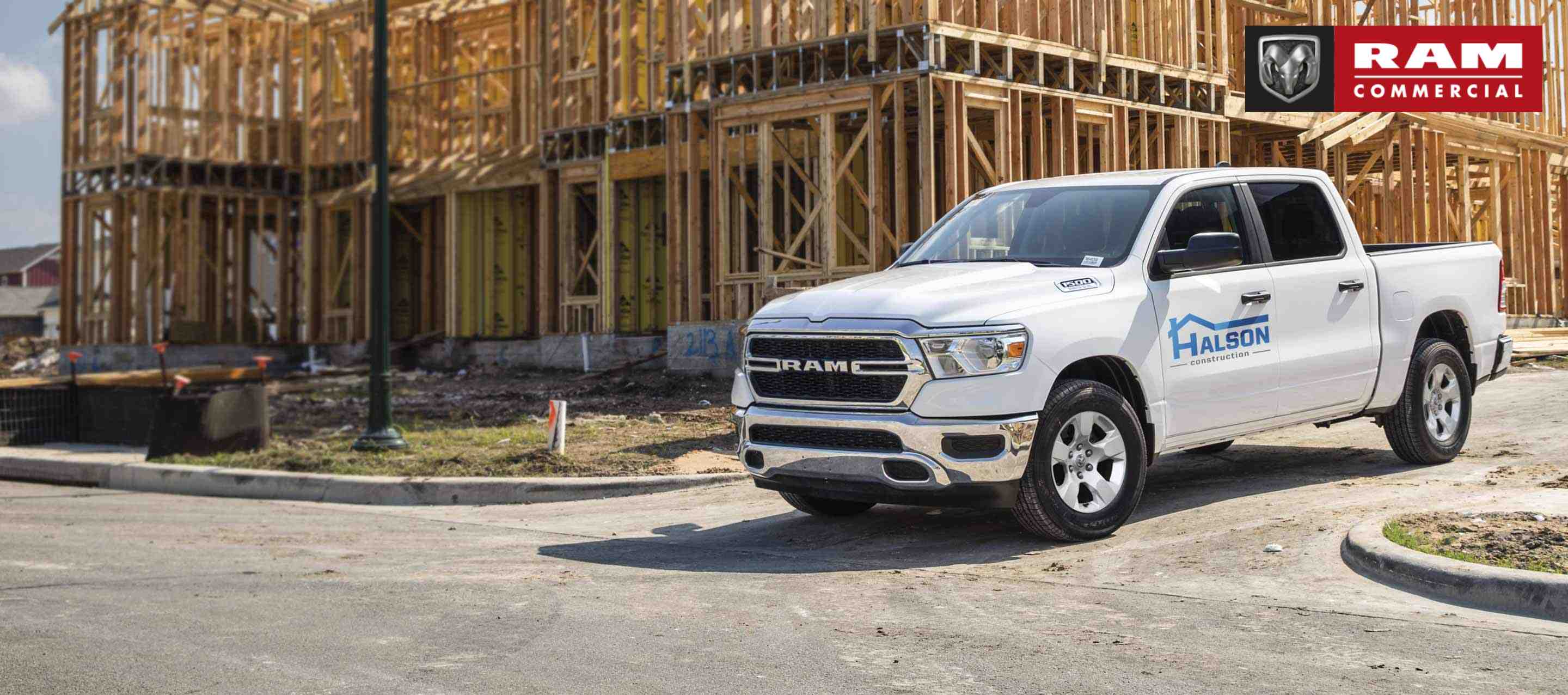 A white 2023 Ram 1500 Tradesman Crew Cab with construction company signage on its driver's door, pulling out of the driveway of a building under construction. Ram Commercial.