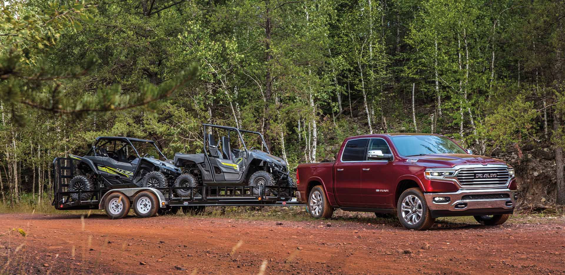 Display The 2023 Ram 1500 parked off-road on a dirt trail with a trailer bearing two ATVs in tow.