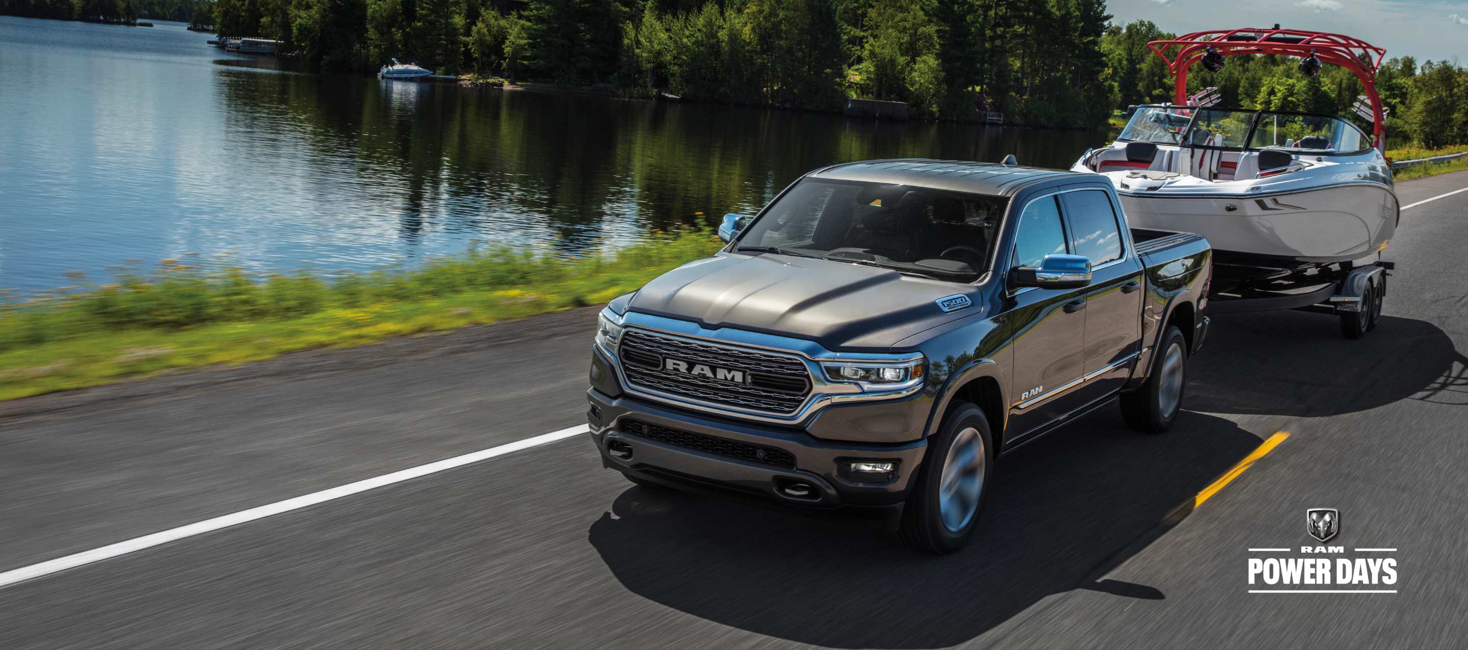 A silver 2023 Ram 1500 Limited 4x4 Crew Cab being driven on a lakeside highway, while towing a motorboat. Power Days Sales Event.