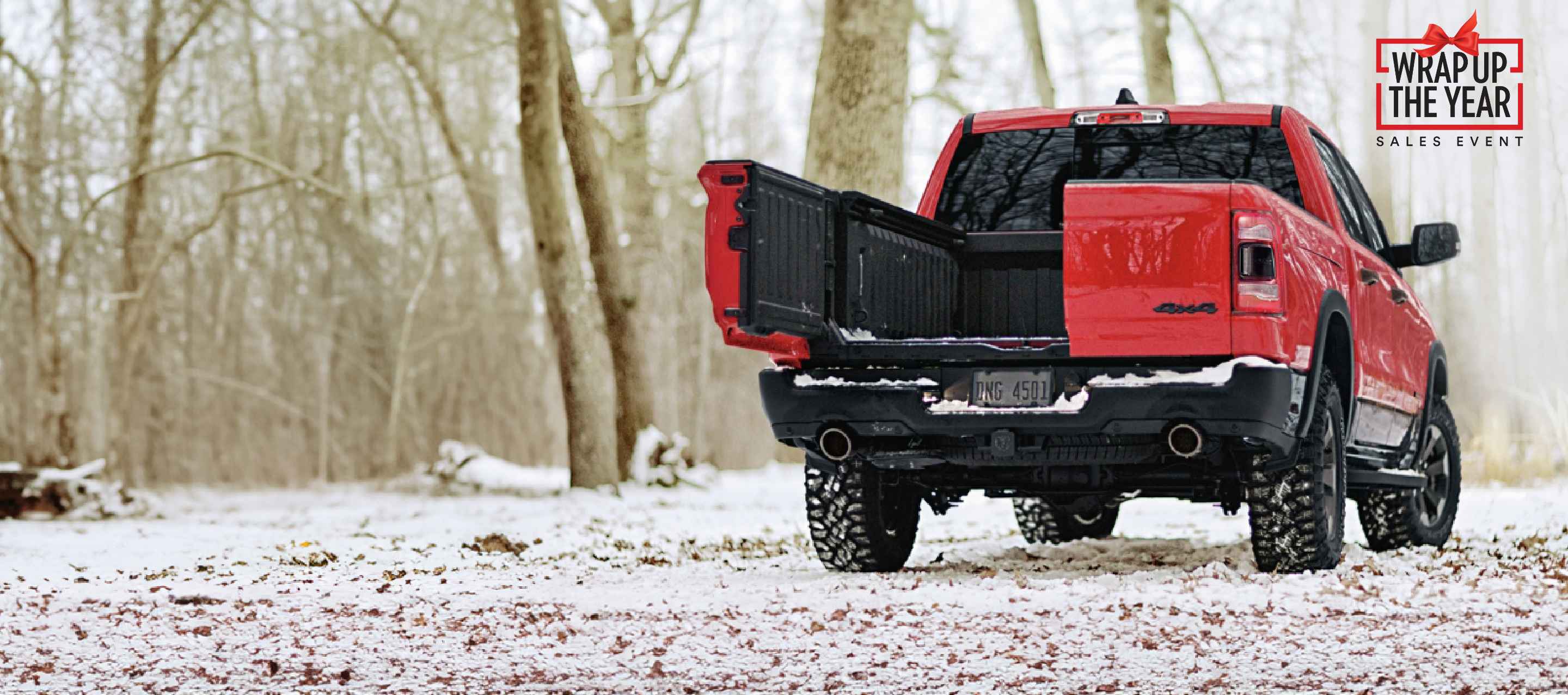 A rear angle of a red 2023 Ram 1500 Rebel 4x4 Crew Cab parked on a snow-covered trail, with one of its multifunction tailgate swing-away doors open. Ram Wrap Up the Year Sales Event.